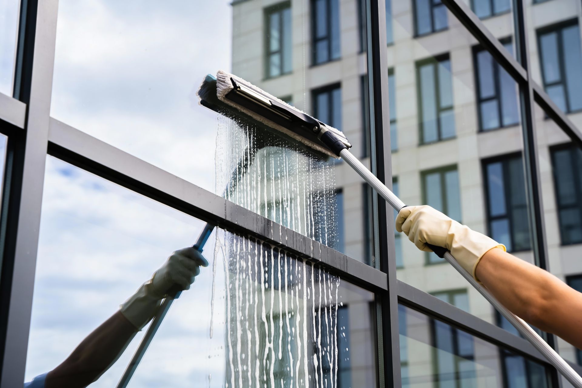 A gloved hand uses a squeegee with soapy water to clean a large glass office building window.