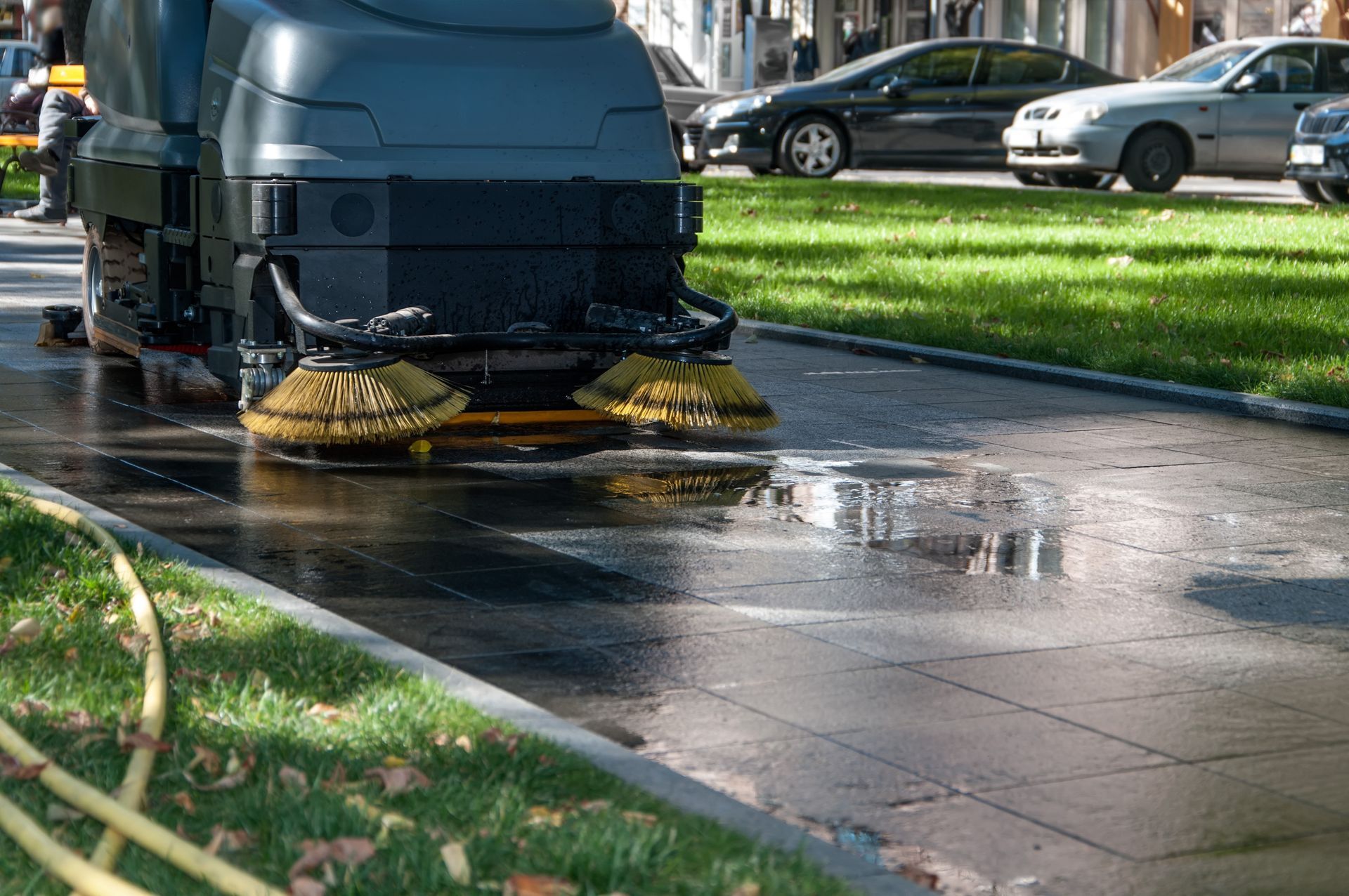 A mechanical sweeper cleans a wet paved walkway bordered by green grass, with parked cars visible in the background.