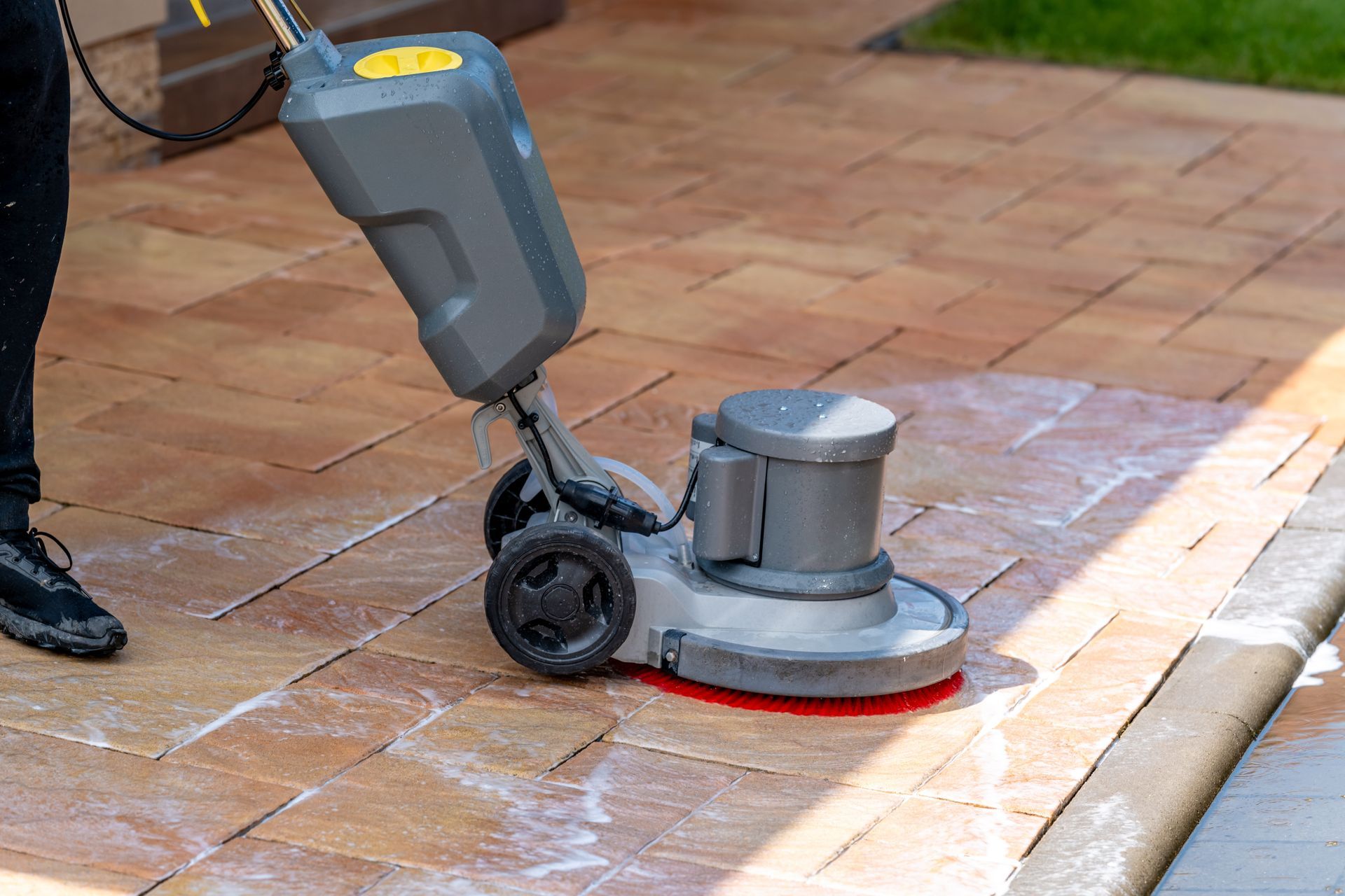A person uses a floor scrubbing machine to clean light brown patio pavers.