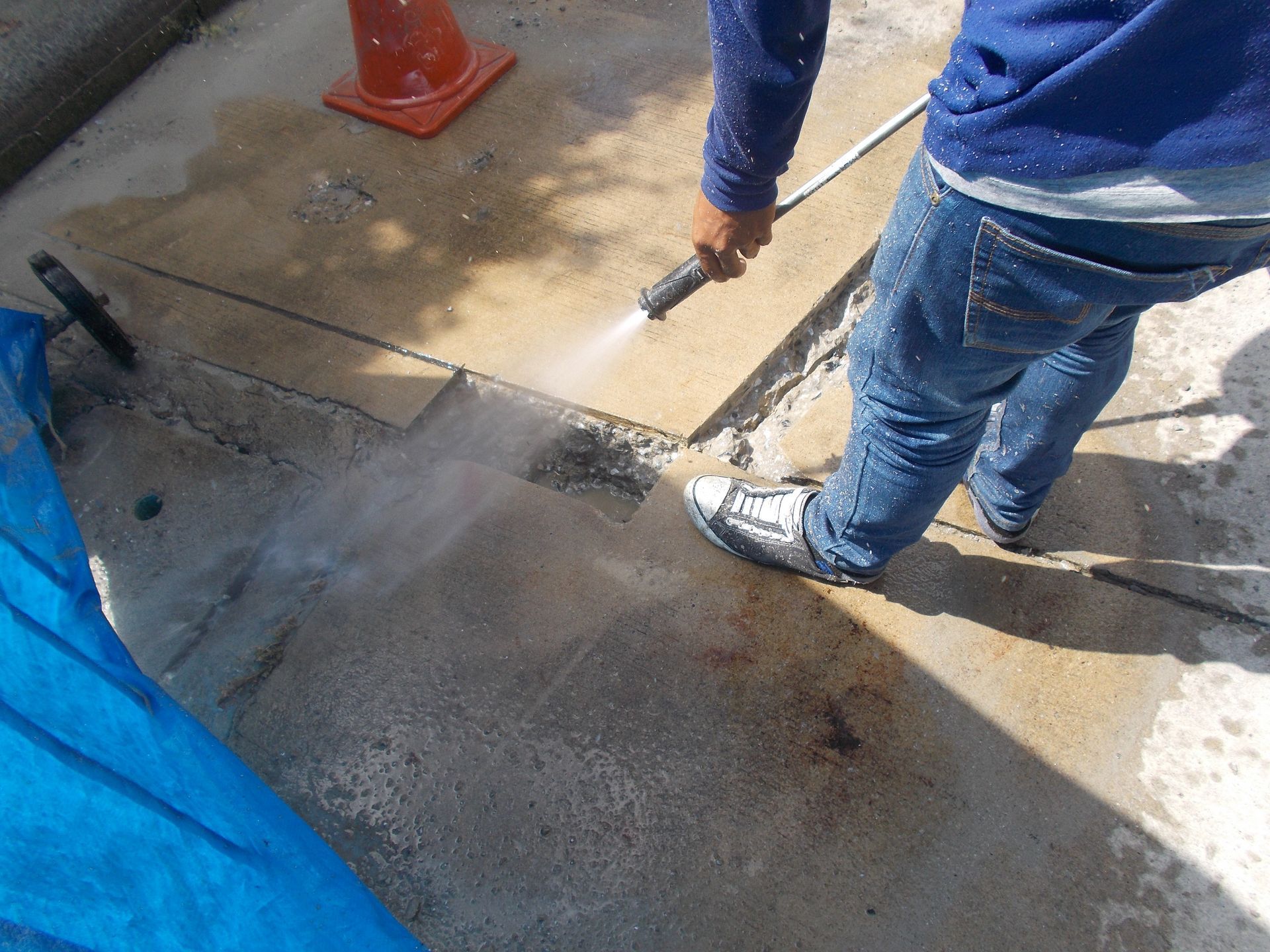 A person in blue jeans and a blue long-sleeved shirt pressure washes a dirty concrete surface outdoors near a traffic cone.