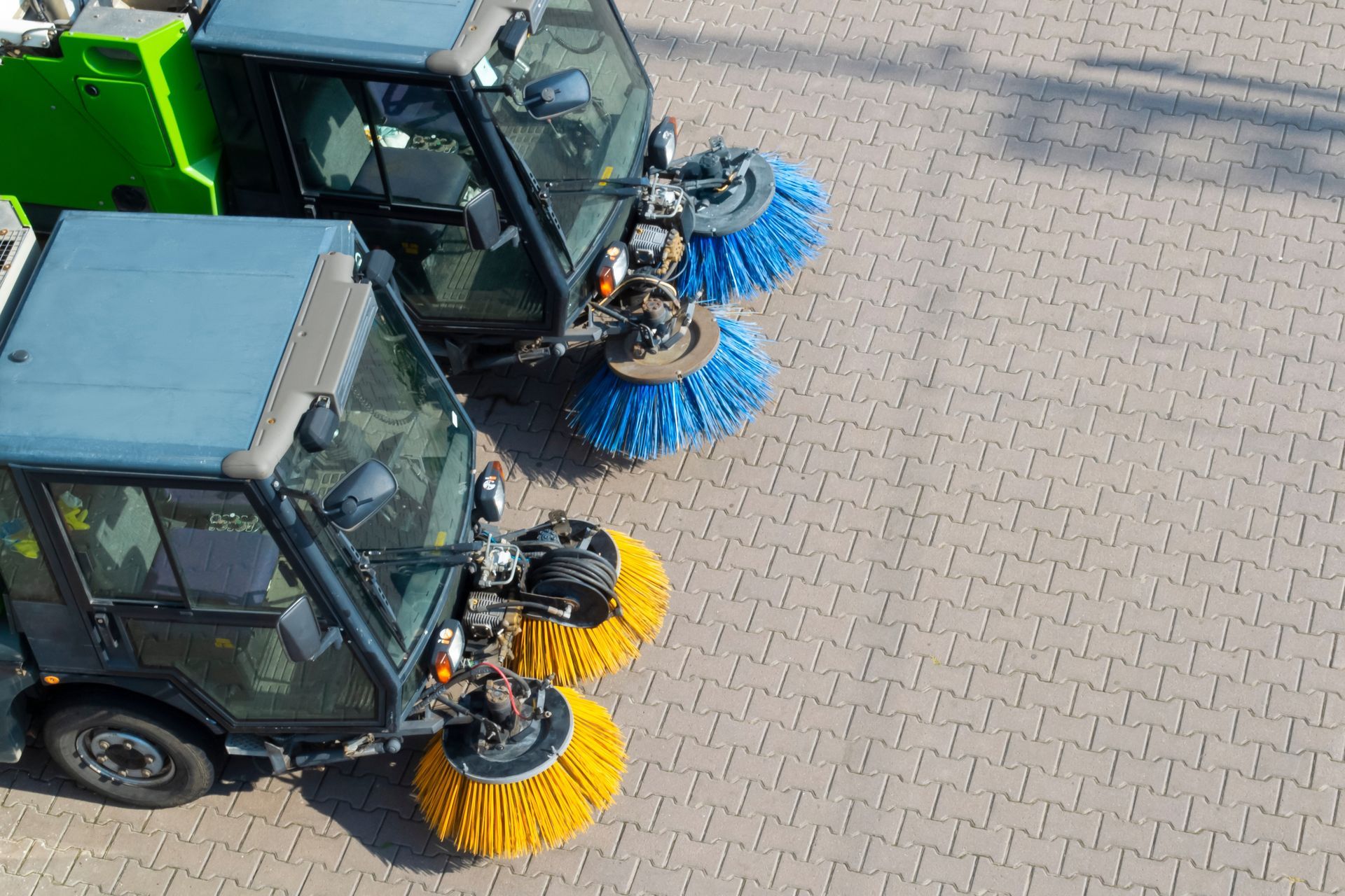 Two street sweepers parked on a paved lot, one with blue bristles and one with yellow bristles.
