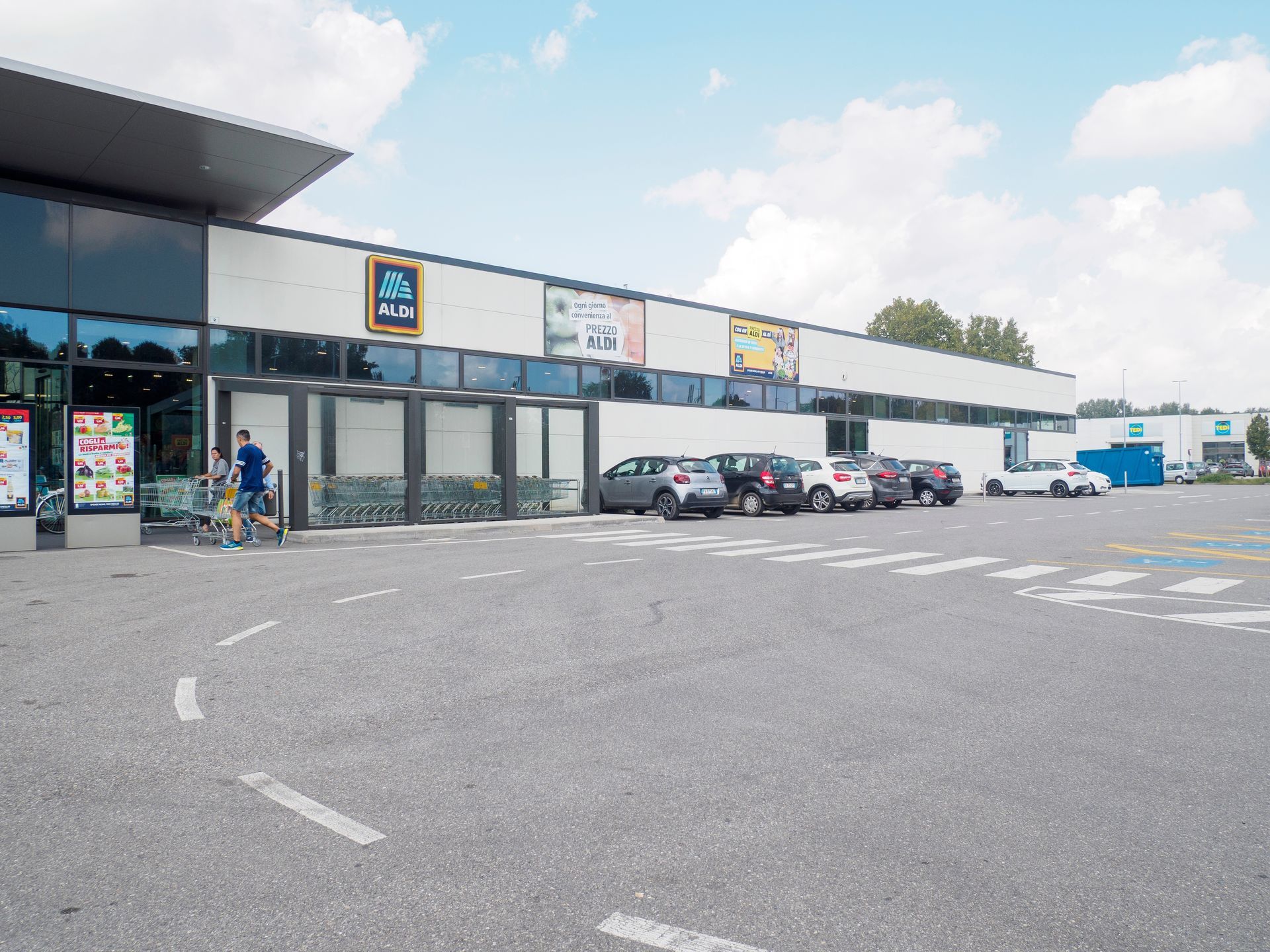 An Aldi supermarket with a gray parking lot and cars in front under a partly cloudy sky.