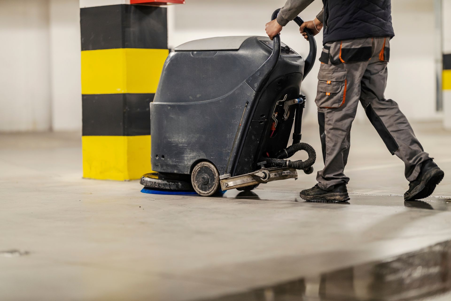 A person operates a floor scrubbing machine in a parking garage next to a yellow and black striped pillar.