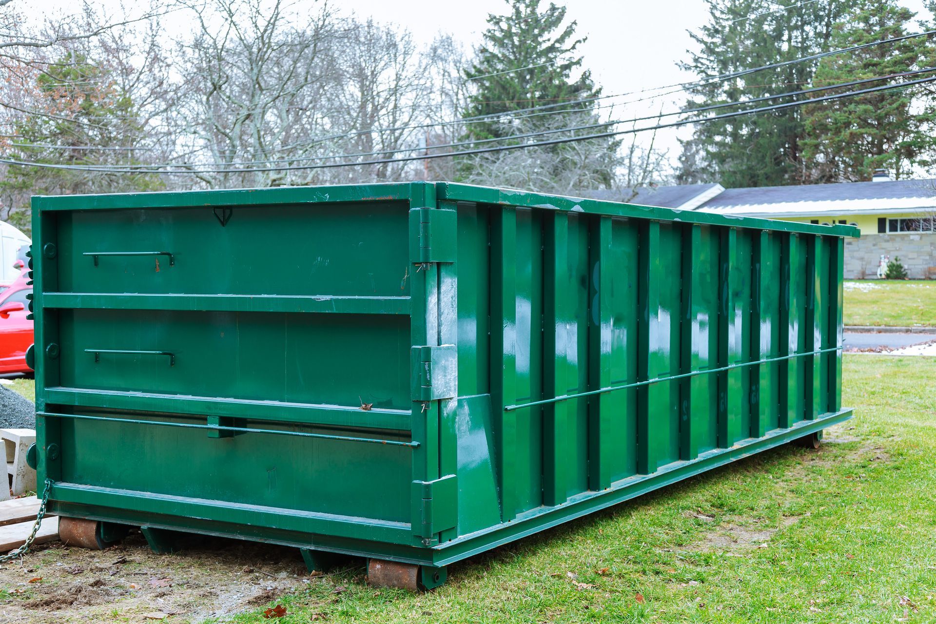 A large, empty rectangular green dumpster sits on a grassy lot in front of a residential house.
