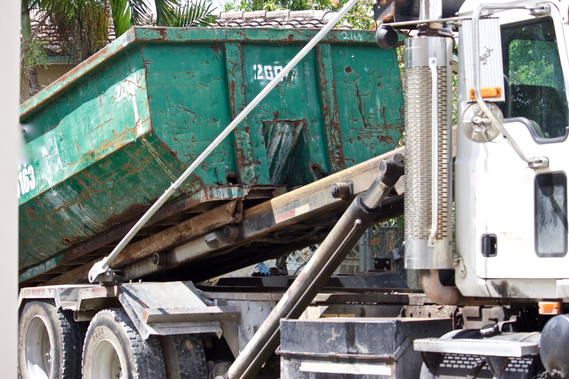 A large green roll-off dumpster being loaded onto the back of a white commercial flatbed truck.