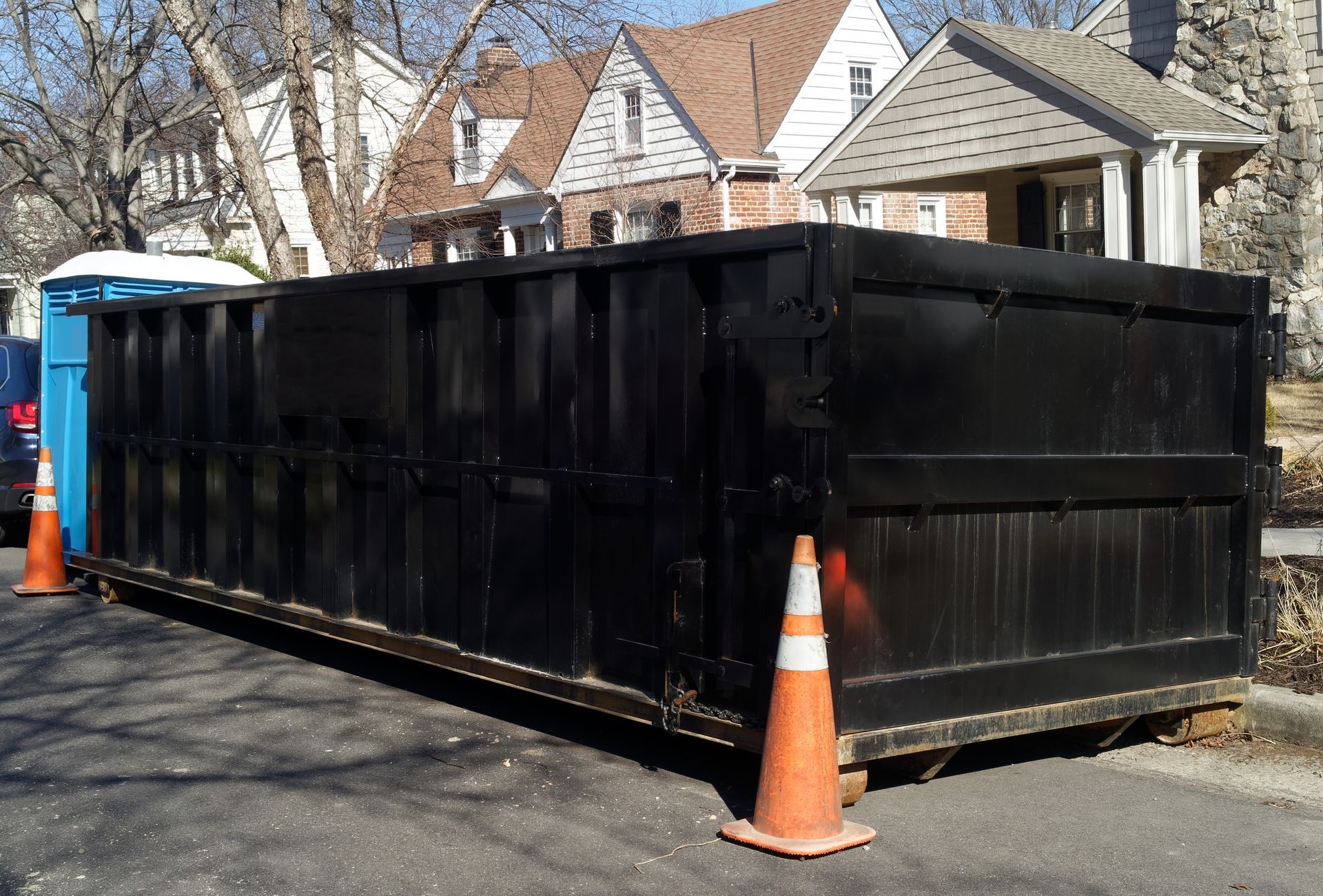 A large, black, rectangular roll-off dumpster sits on a paved street in front of suburban houses with orange traffic cones.