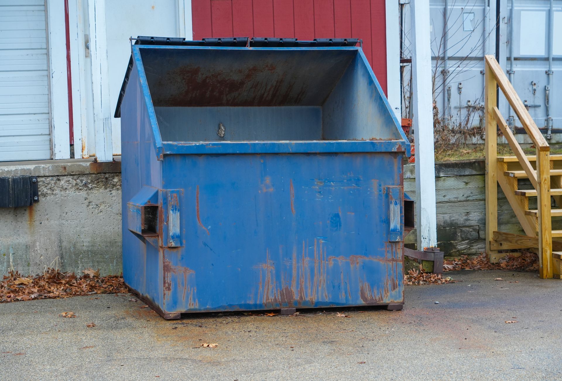 A blue, weathered industrial dumpster sits on pavement next to a red-walled building with wooden stairs.
