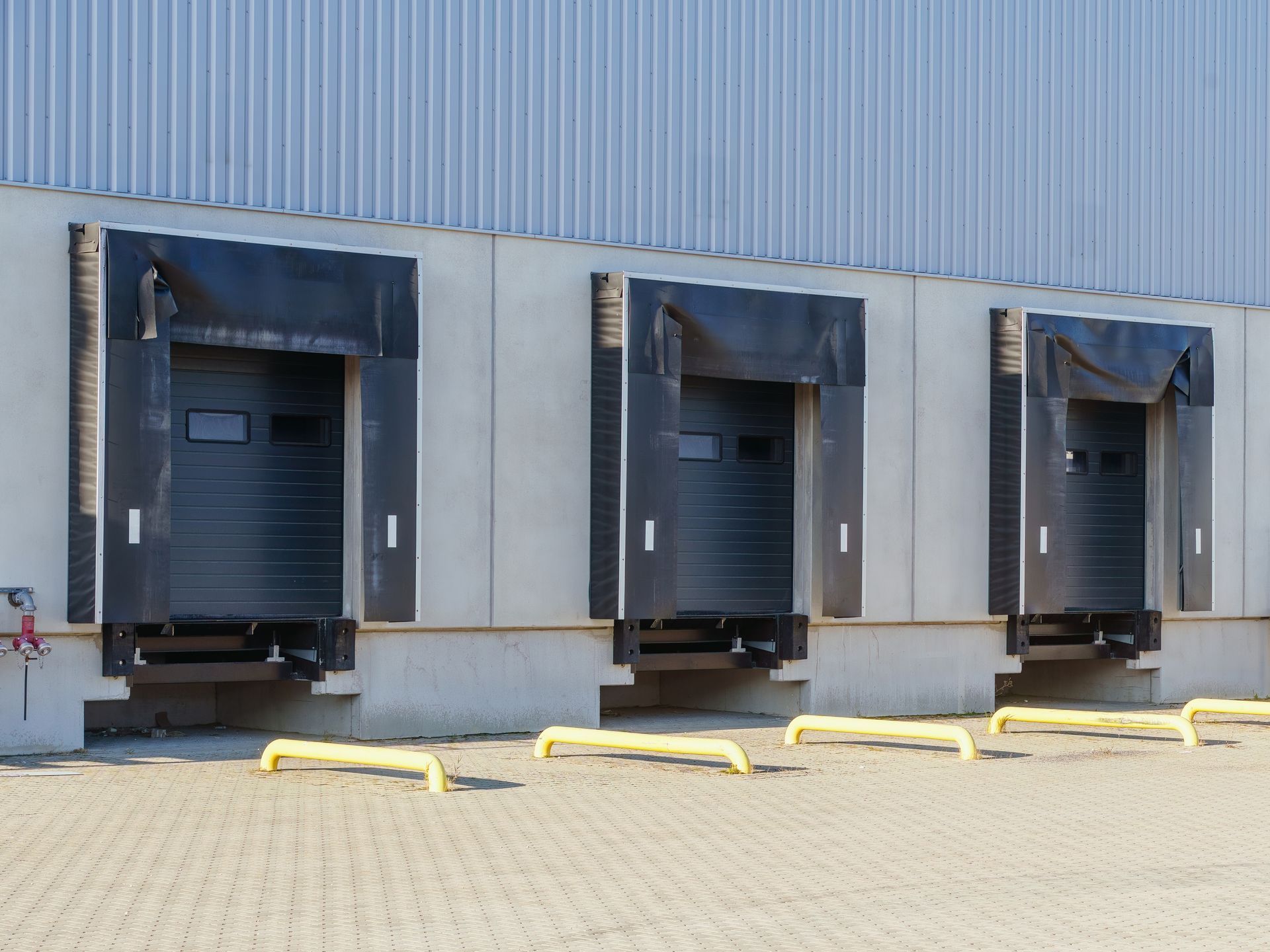 Three industrial loading dock bays with black doors and dock shelters on a concrete wall above a paved yard.