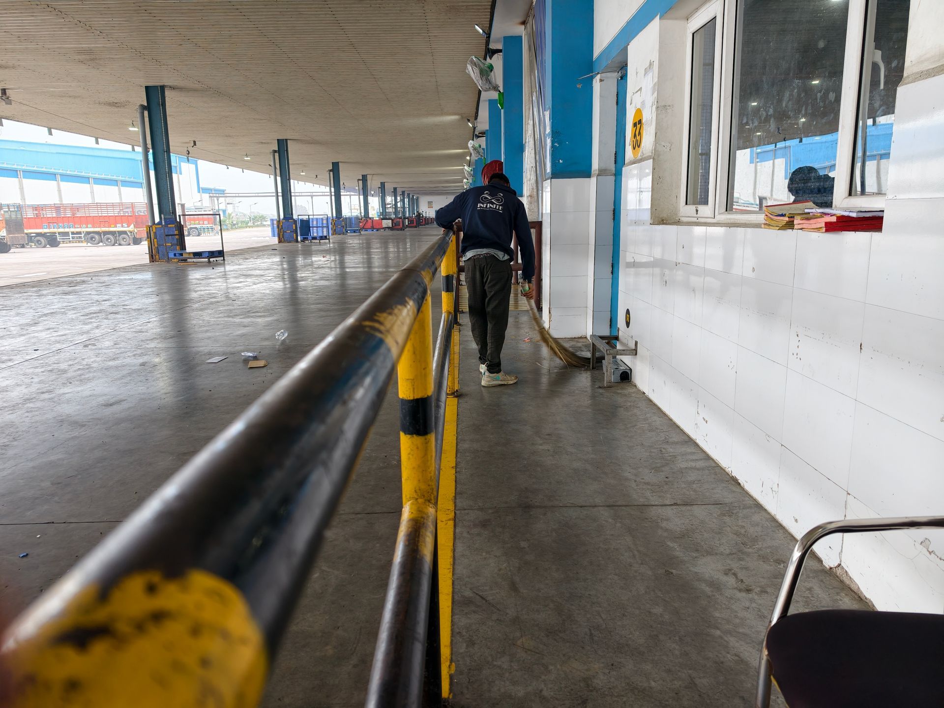 A person in dark work clothes sweeps a concrete floor alongside a yellow and black safety railing at a train station.