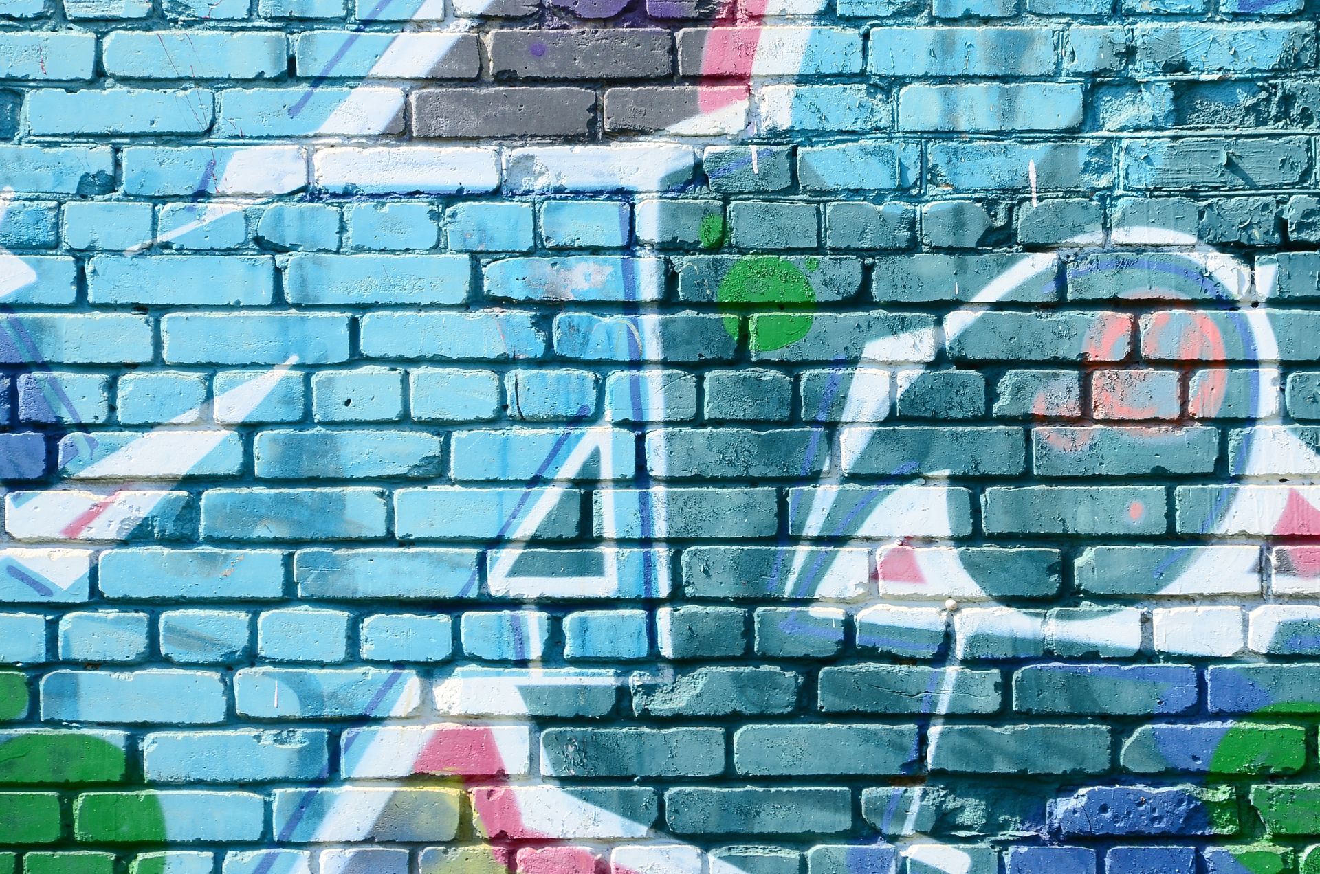 A close-up of a blue brick wall covered in layered white, pink, and green graffiti patterns.