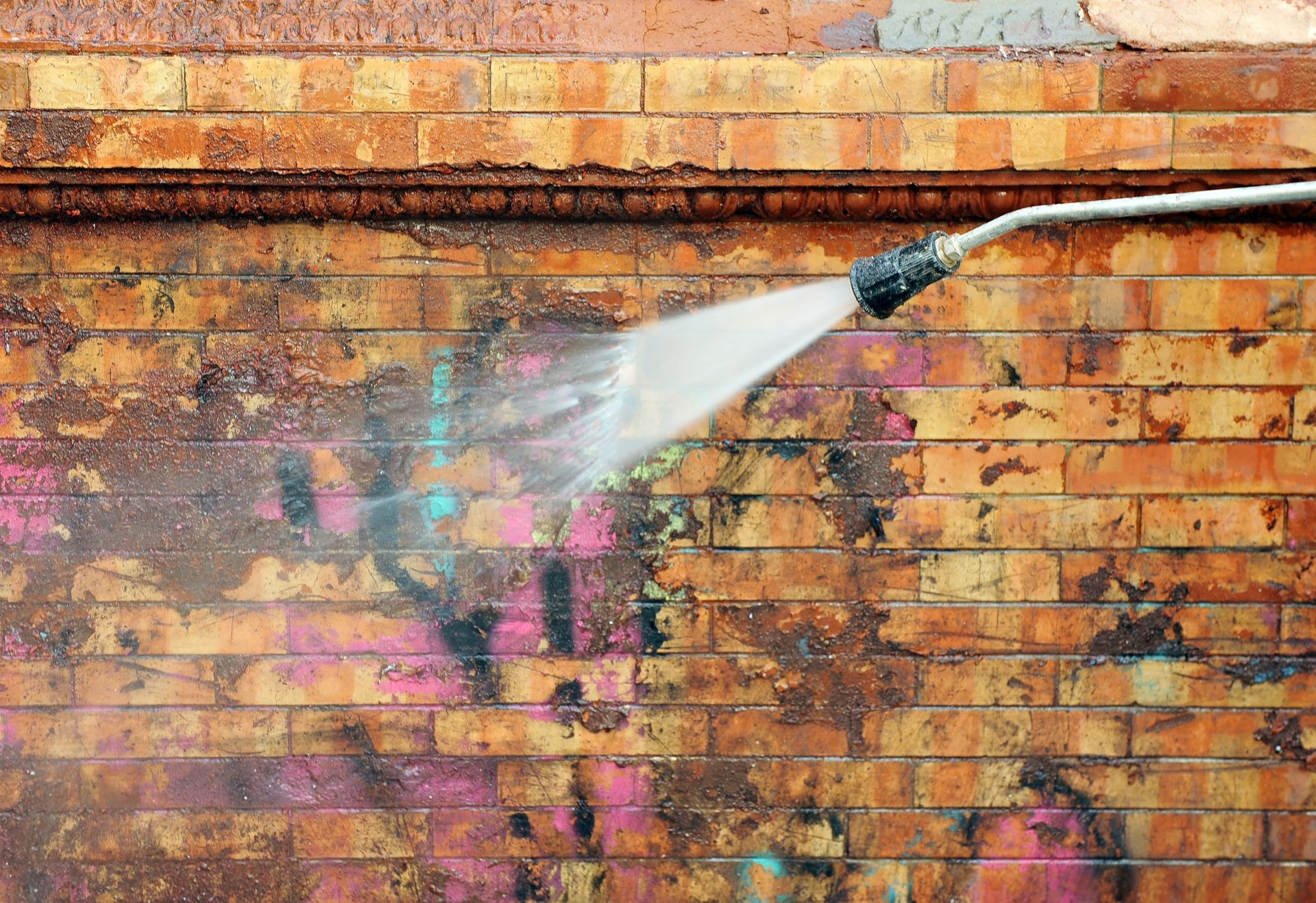 A pressure washer sprays water on a stained, multi-colored brick wall to clean off grime and paint.