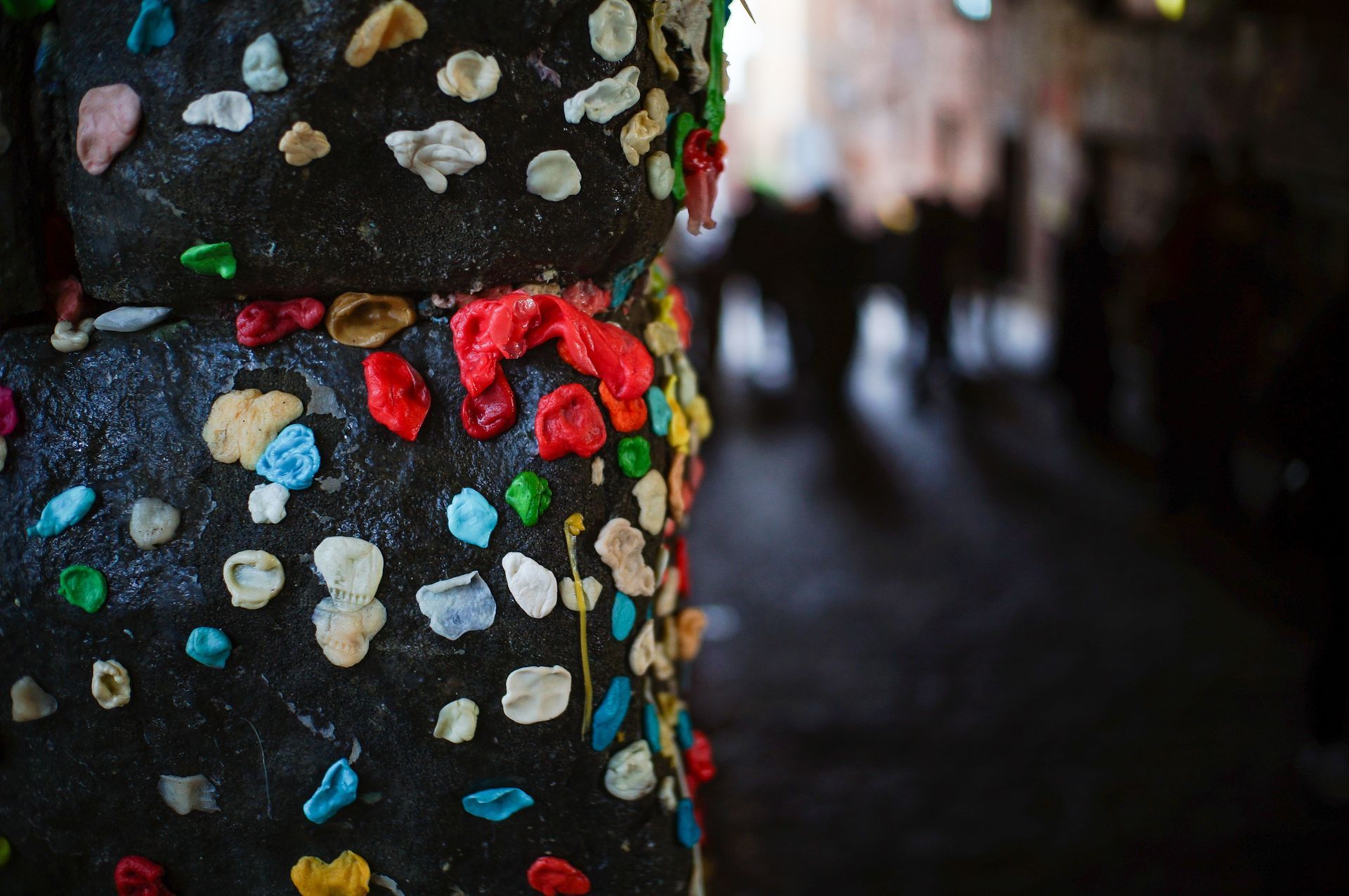 A close-up of a dark, textured wall covered in colorful pieces of discarded chewing gum, with a blurred street background.