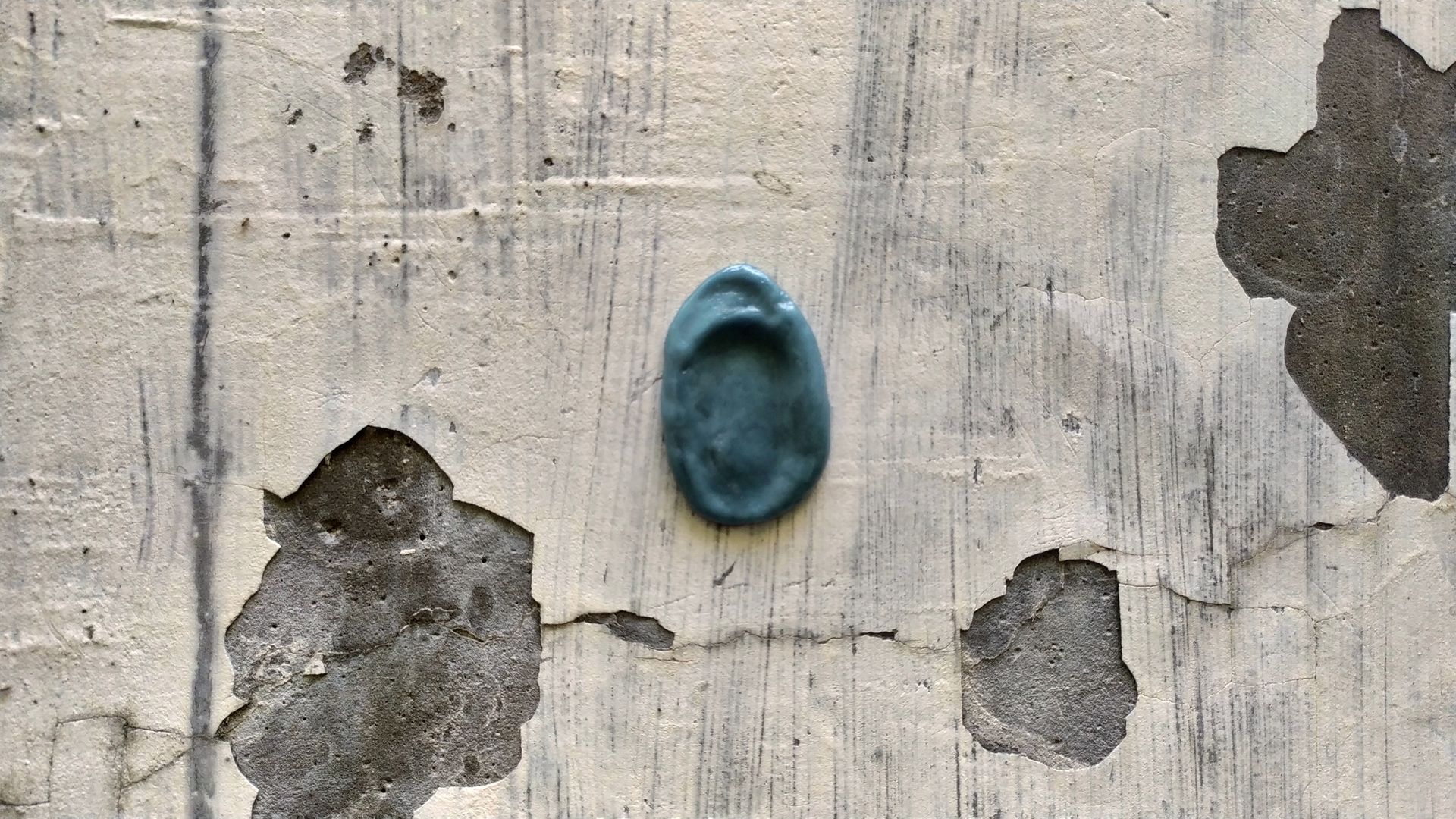 A small, teal, ear-shaped sculpture is mounted on a weathered, light-colored stone wall with patches of exposed concrete.