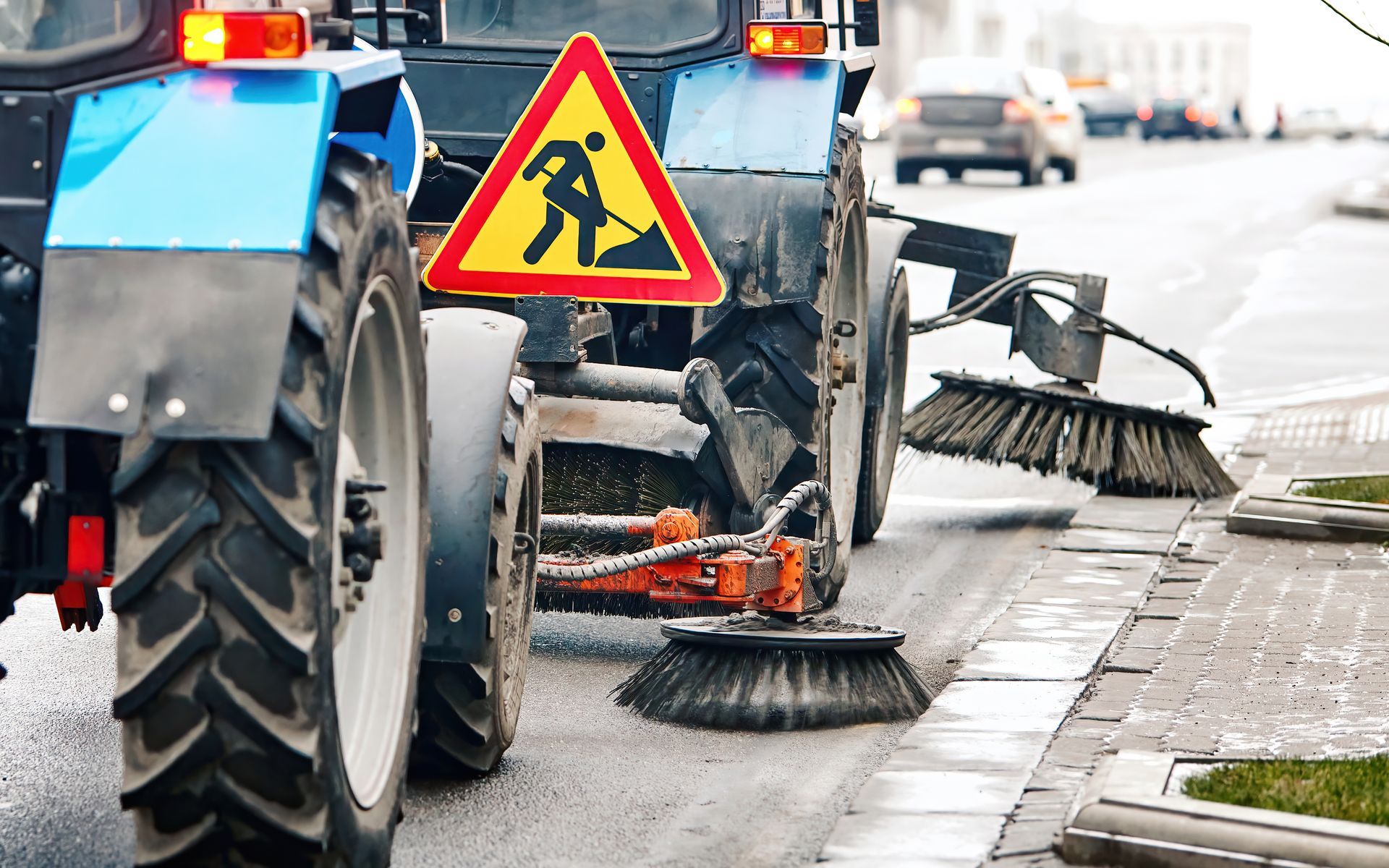 A blue street sweeper tractor with a yellow construction warning sign works on a city road.