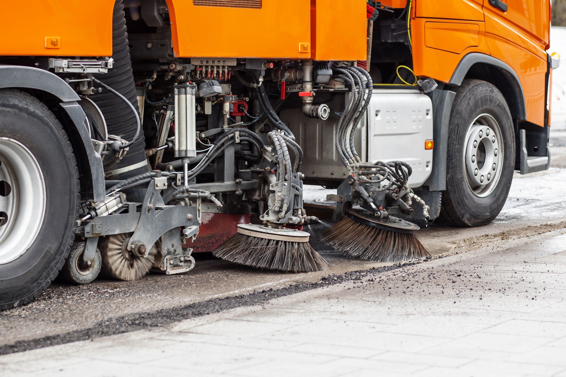 An orange street sweeper truck with rotating brushes cleaning debris along the edge of a paved road.