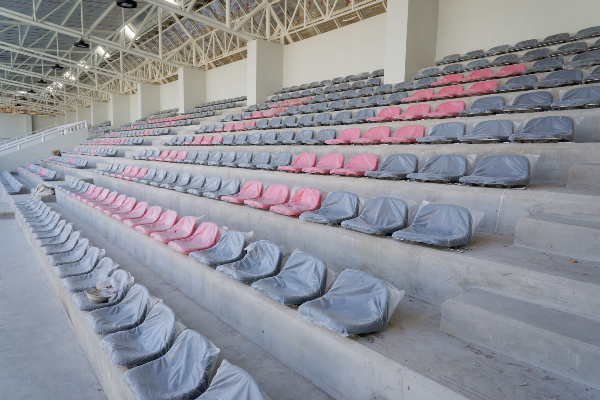 An indoor stadium arena featuring rows of gray and pink plastic seats on tiered concrete steps under a metal roof.