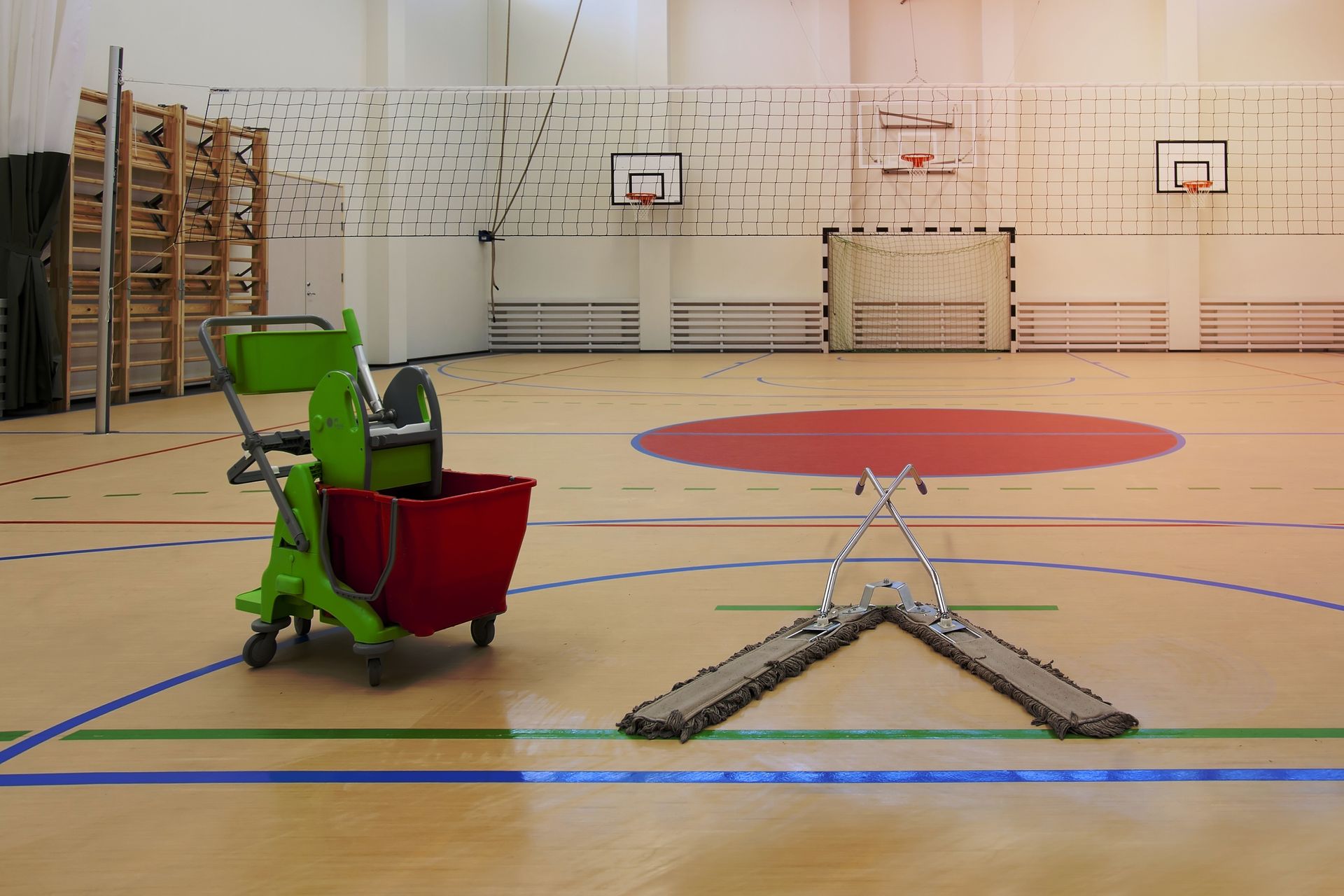 A green mop bucket cart and a V-shaped floor sweeper sit on the light-colored hardwood floor of an indoor sports gym.