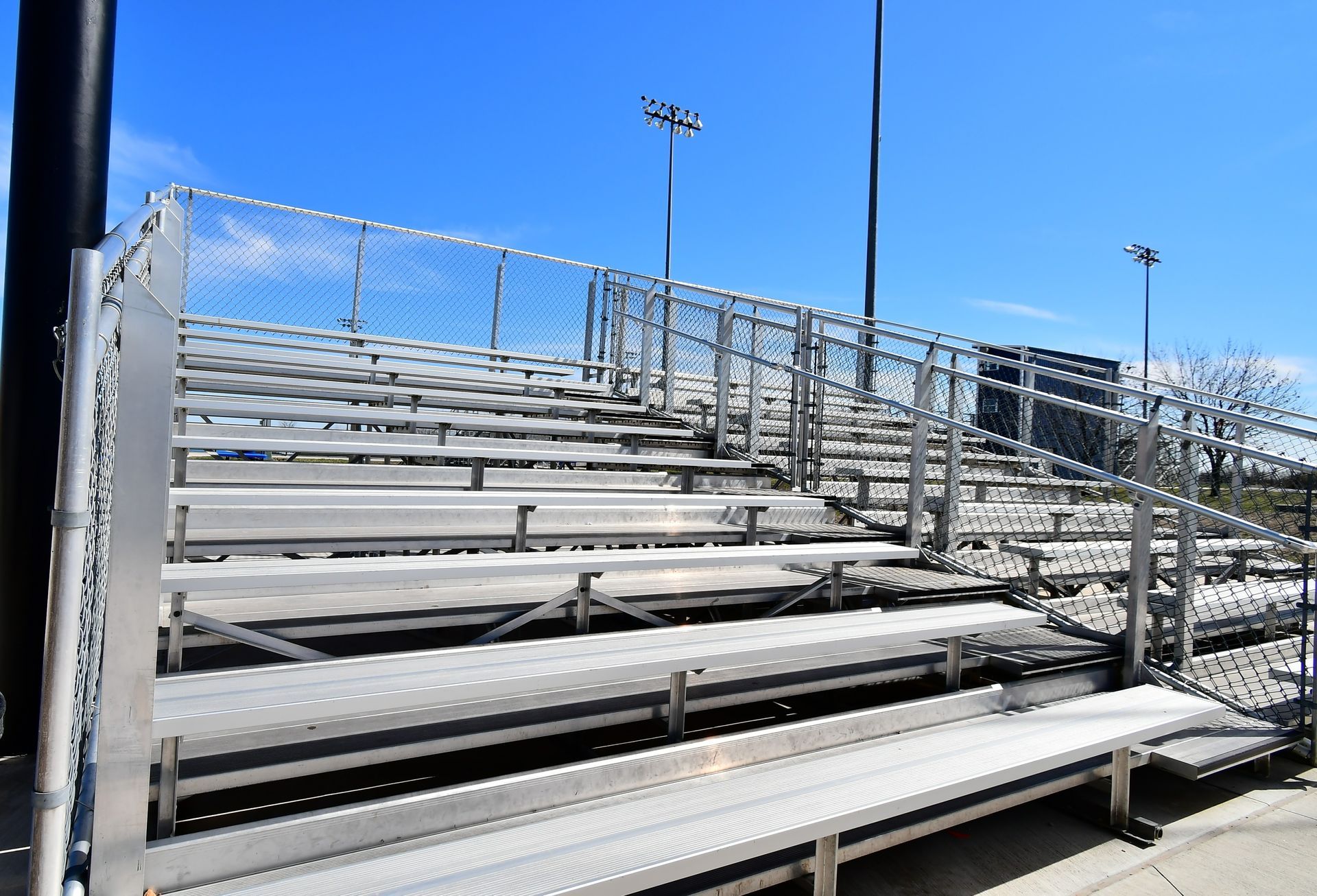 Empty tiered metal bleachers under a bright blue sky with light poles and a chain-link fence.