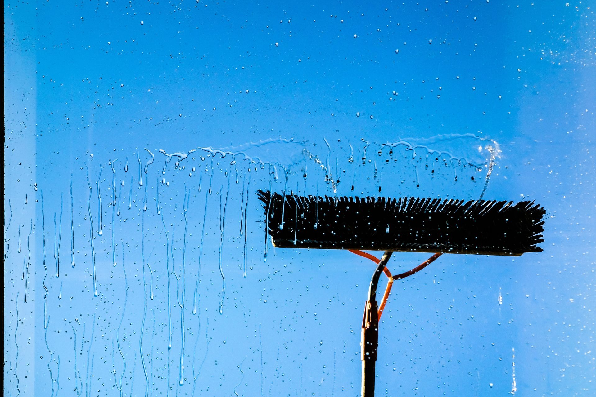 A brush on a pole cleaning a window with water droplets and streaks against a bright blue sky.