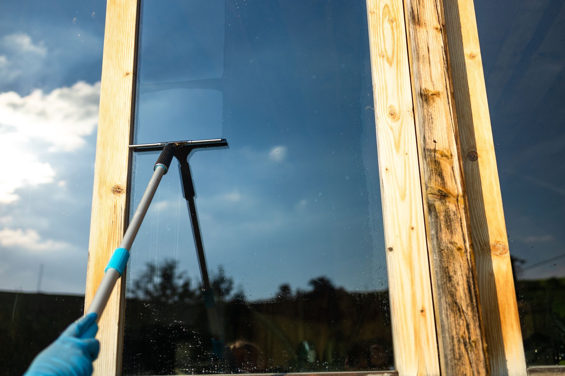 A gloved hand uses a squeegee to clean a tall window framed by natural wood against a bright, cloudy sky.