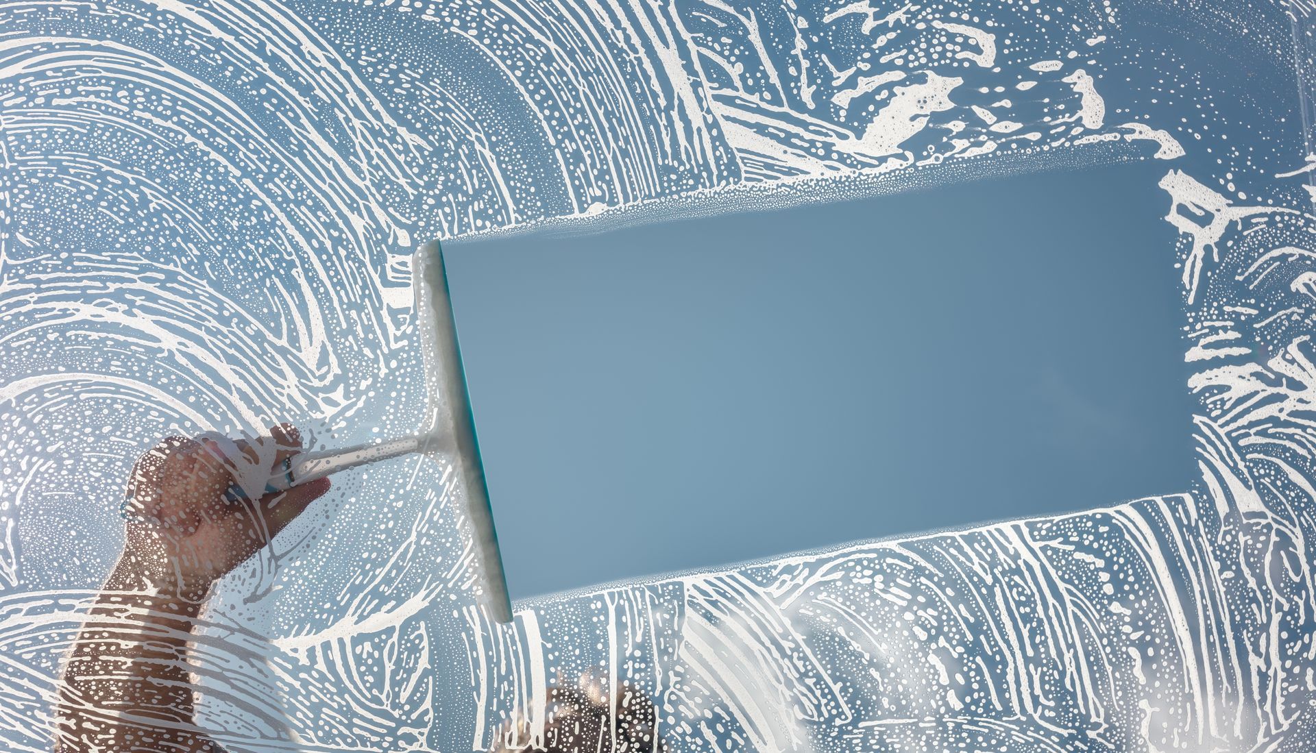 A squeegee wipes away soapy water from a glass pane, revealing a clear blue patch against a sudsy background.
