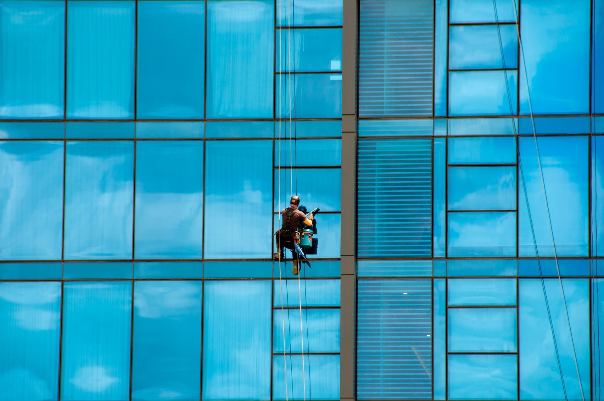 A window washer suspended by ropes cleans the glass exterior of a modern, blue-tinted skyscraper.