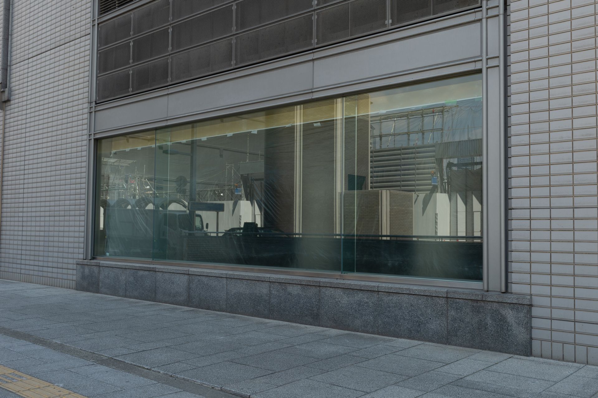 Large, rectangular storefront window on a gray brick building at street level, showing faint interior reflections.