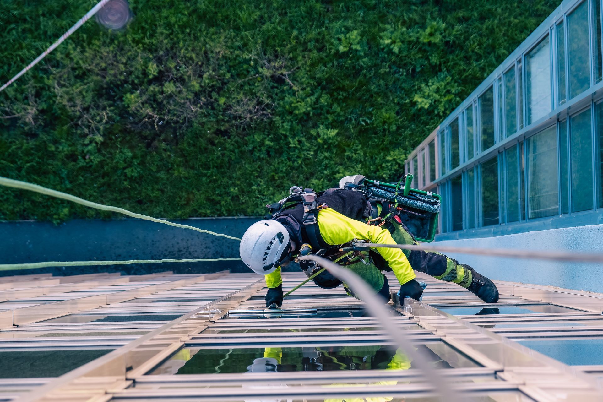 A high-angle view of a worker in a safety helmet and yellow jacket rappelling down the side of a glass building.