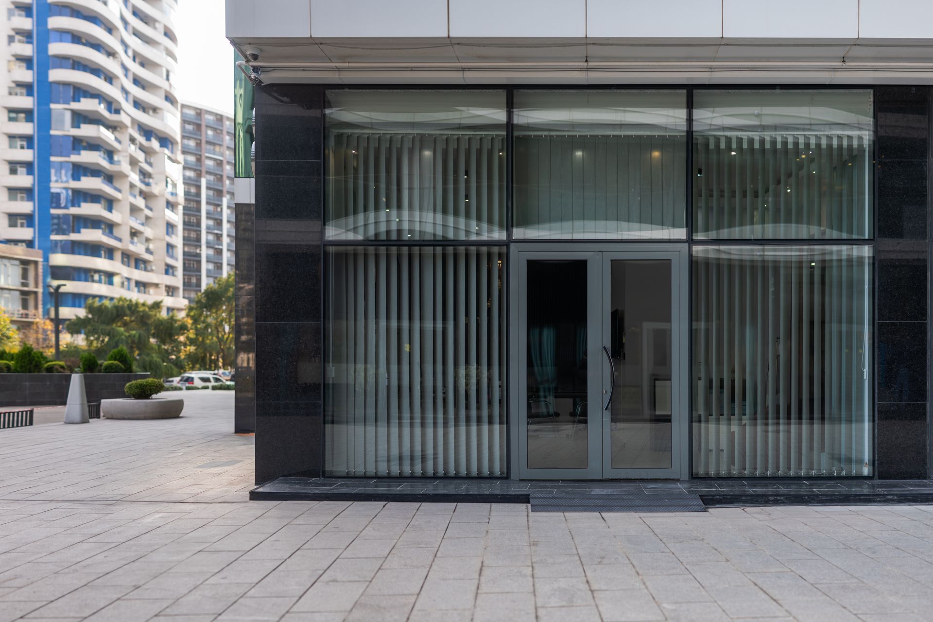 Ground-level view of a modern glass storefront with dark stone cladding and vertical blinds, beside a paved plaza.
