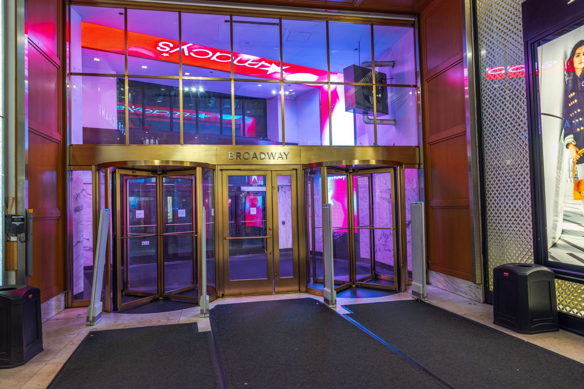 An entrance to a building on Broadway, featuring gold-toned frames, two revolving doors, and neon red and purple lights.