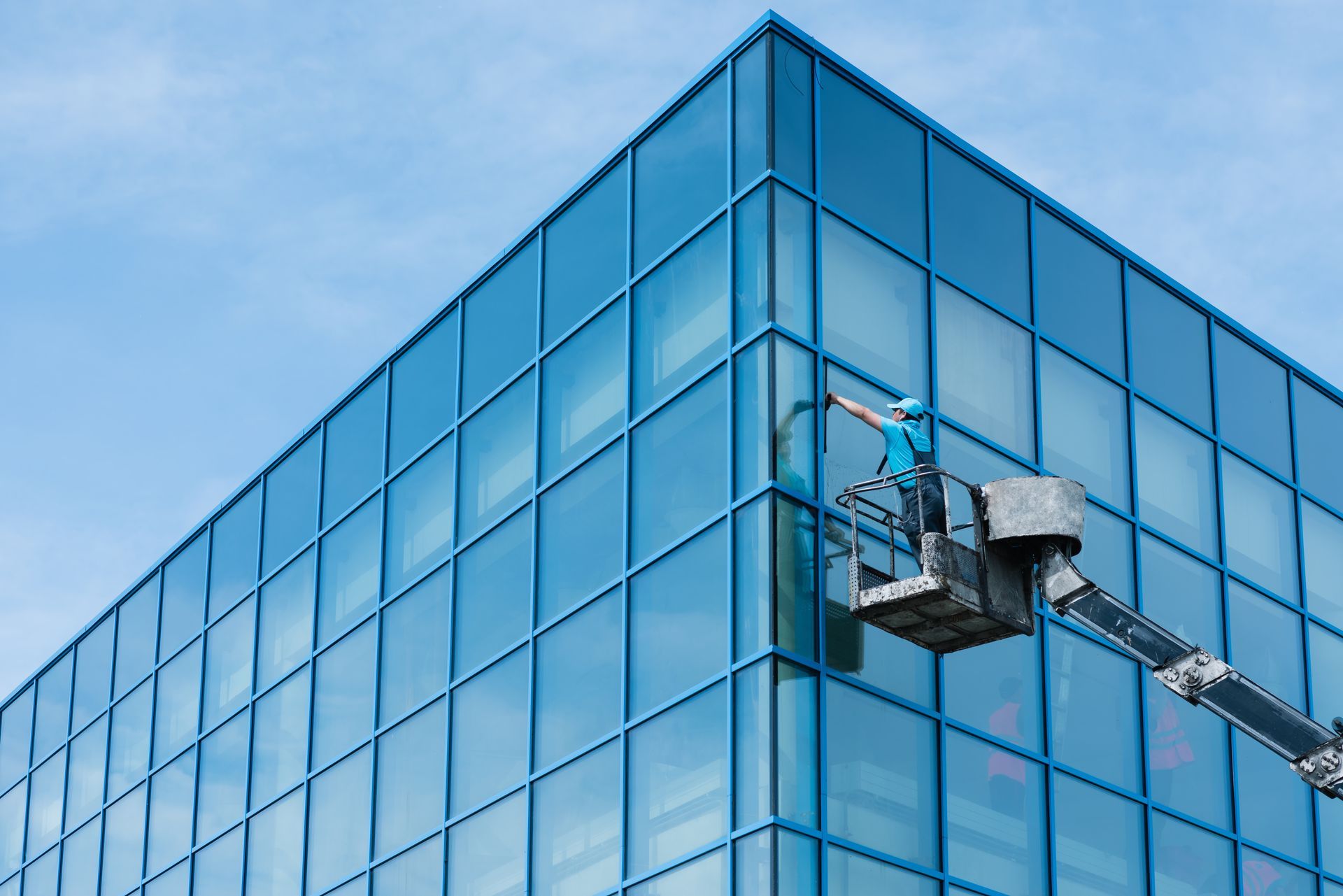 A person in a blue shirt cleans the windows of a modern blue-glass building from an elevated work platform.