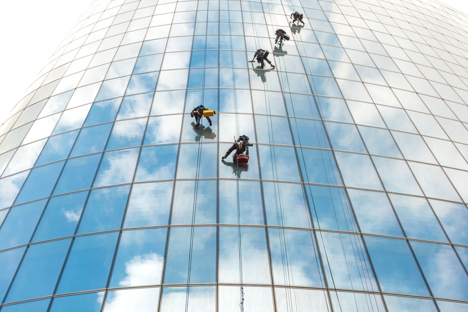 Window washers in safety harnesses descend the side of a modern glass skyscraper reflecting a blue sky with clouds.