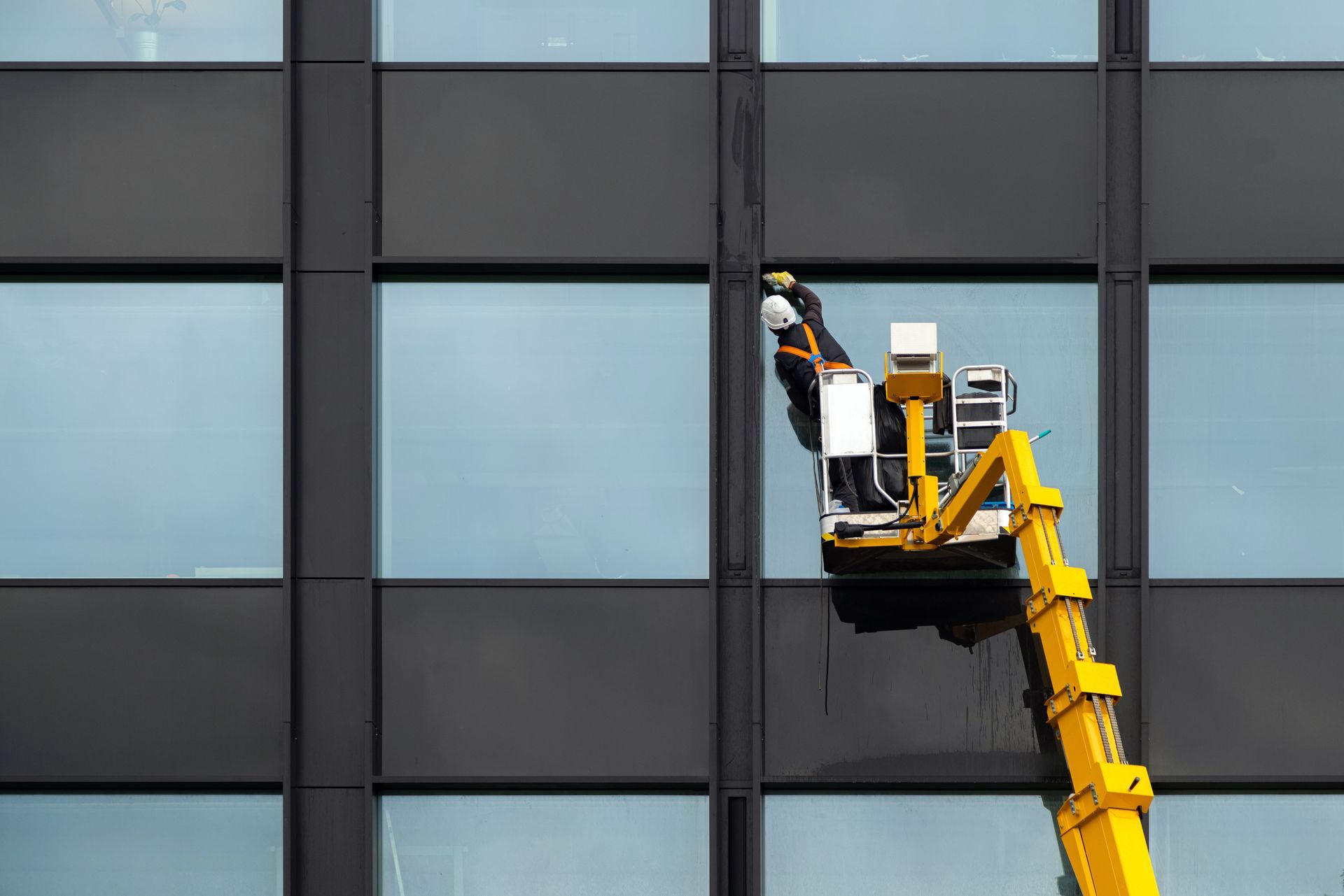 A worker in a safety harness stands in a yellow aerial lift, cleaning the windows of a modern office building.