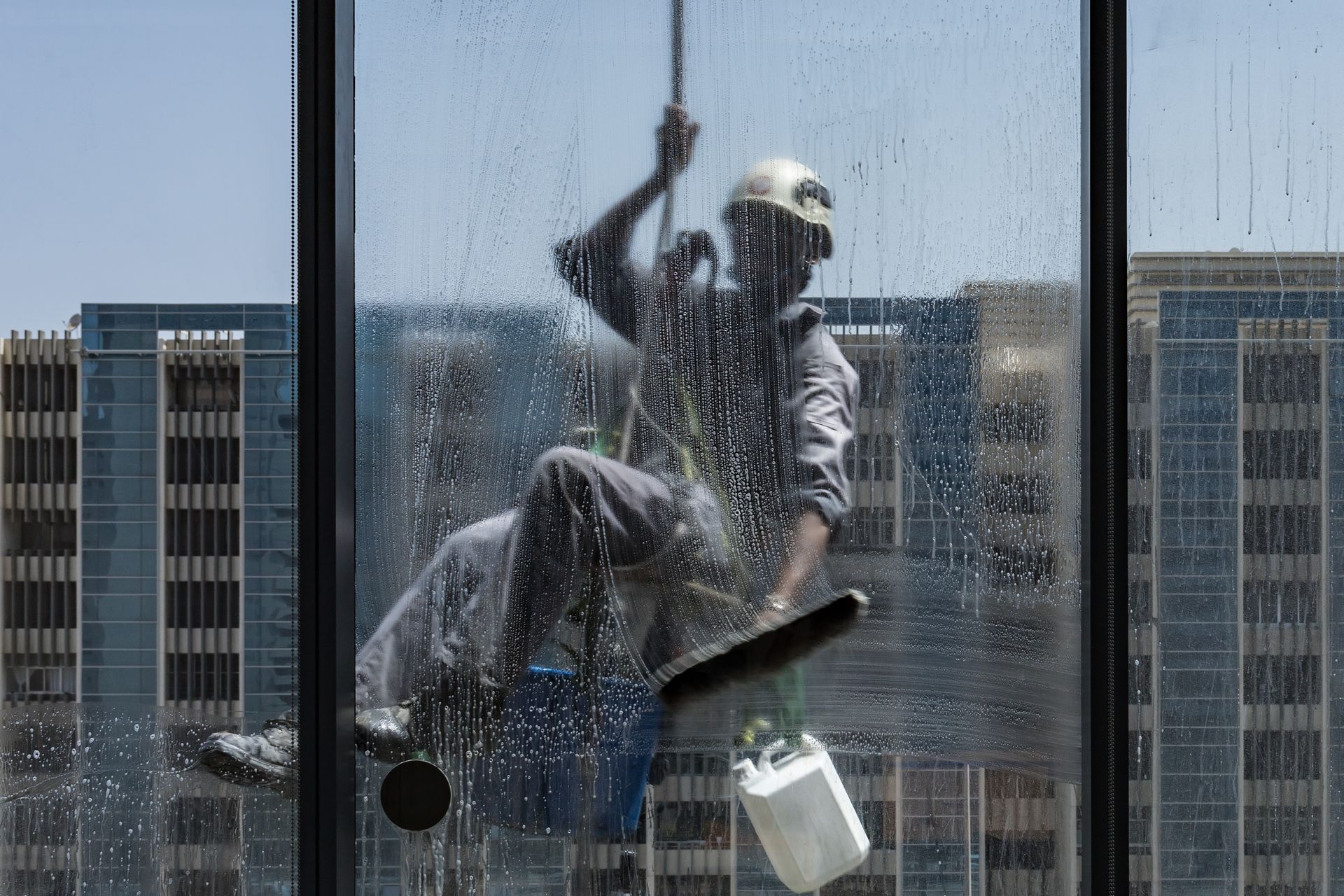 A worker suspended by ropes cleans a large glass window, with city buildings visible in the background.