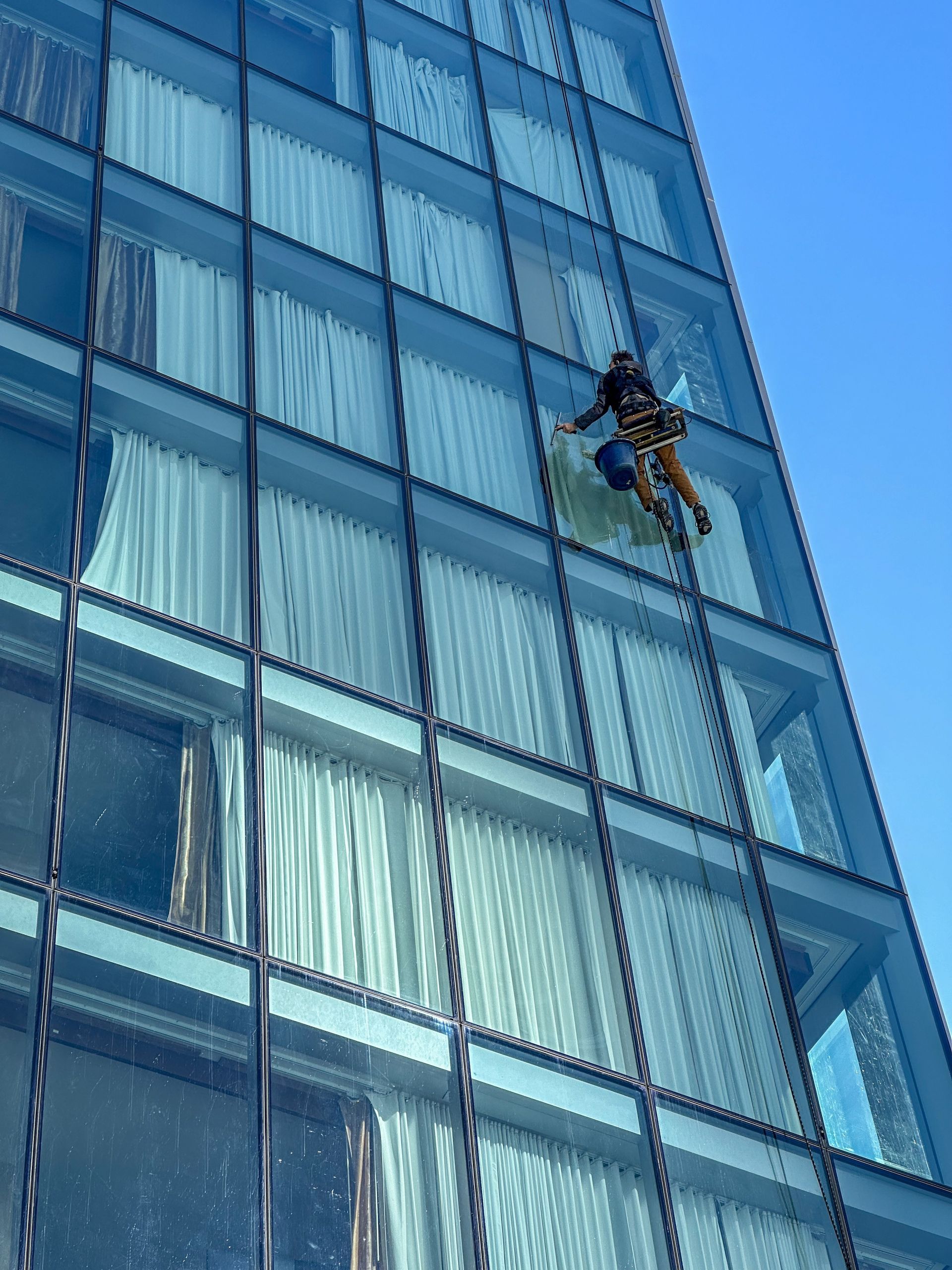 A window cleaner rappelling down the side of a modern glass skyscraper against a clear blue sky.