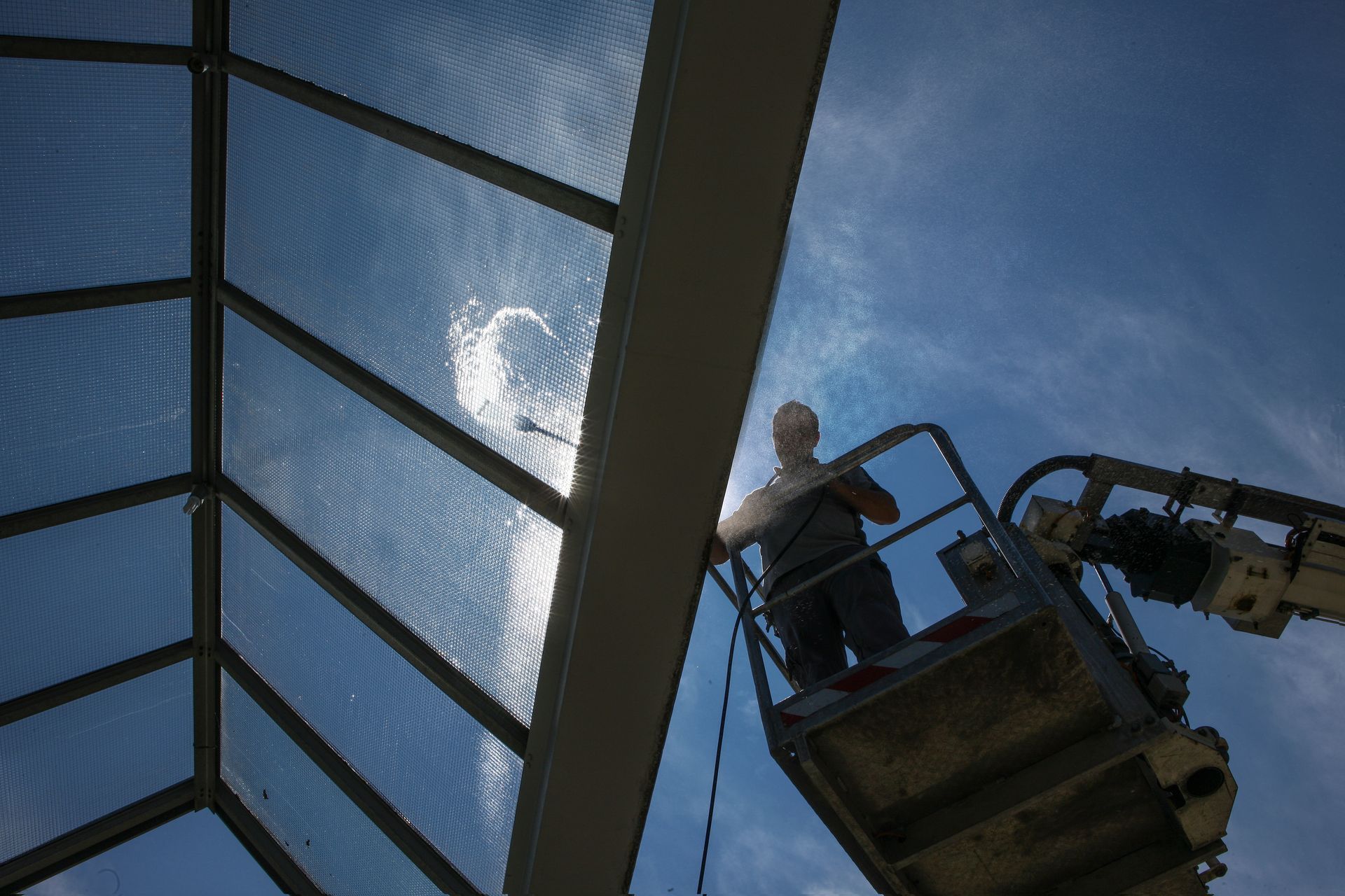 A person in a hydraulic lift uses a long pole to clean a large, framed glass skylight against a bright blue sky.