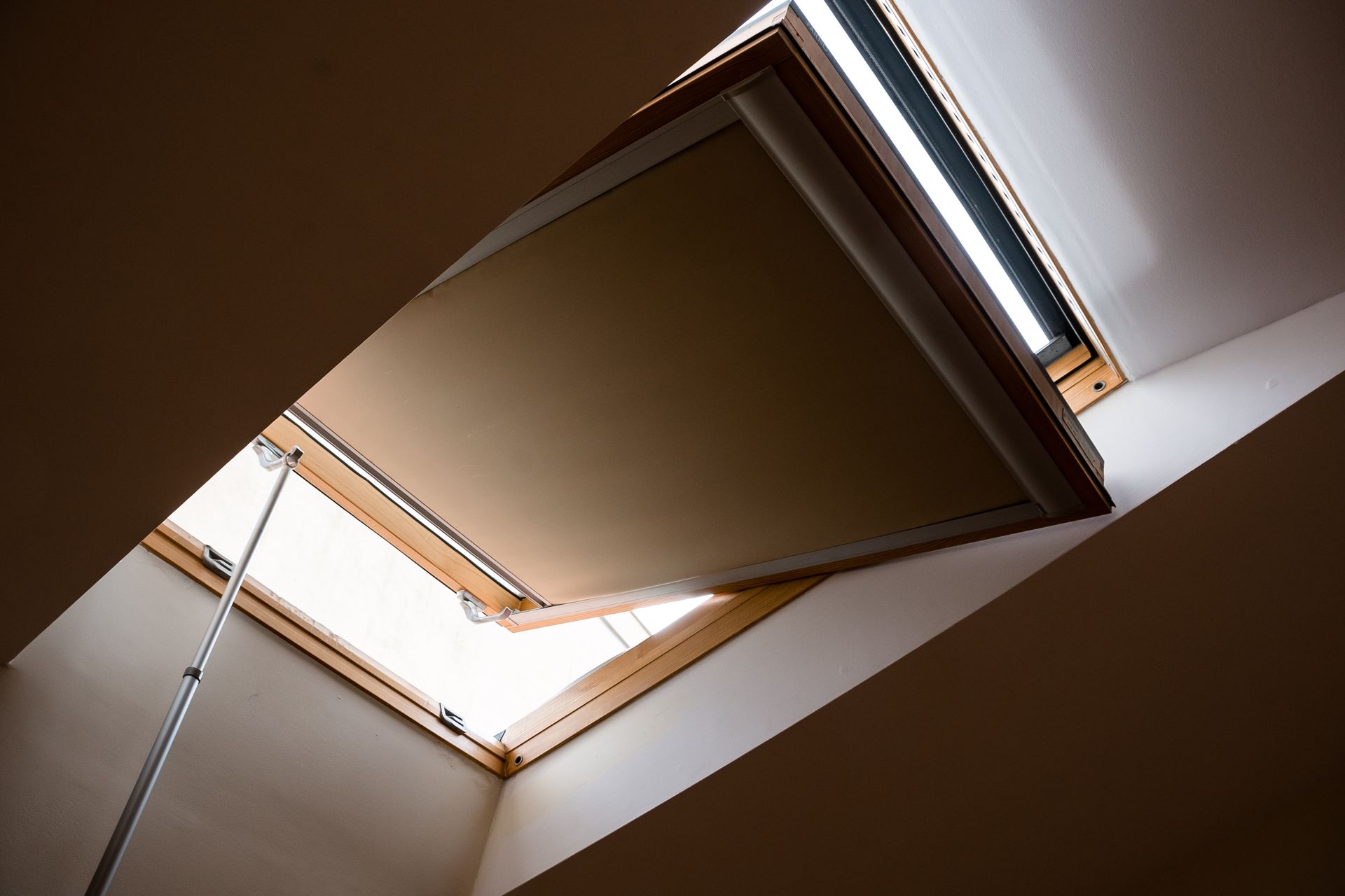 A wooden skylight window tilted open to the sky, viewed from inside a room with white walls.
