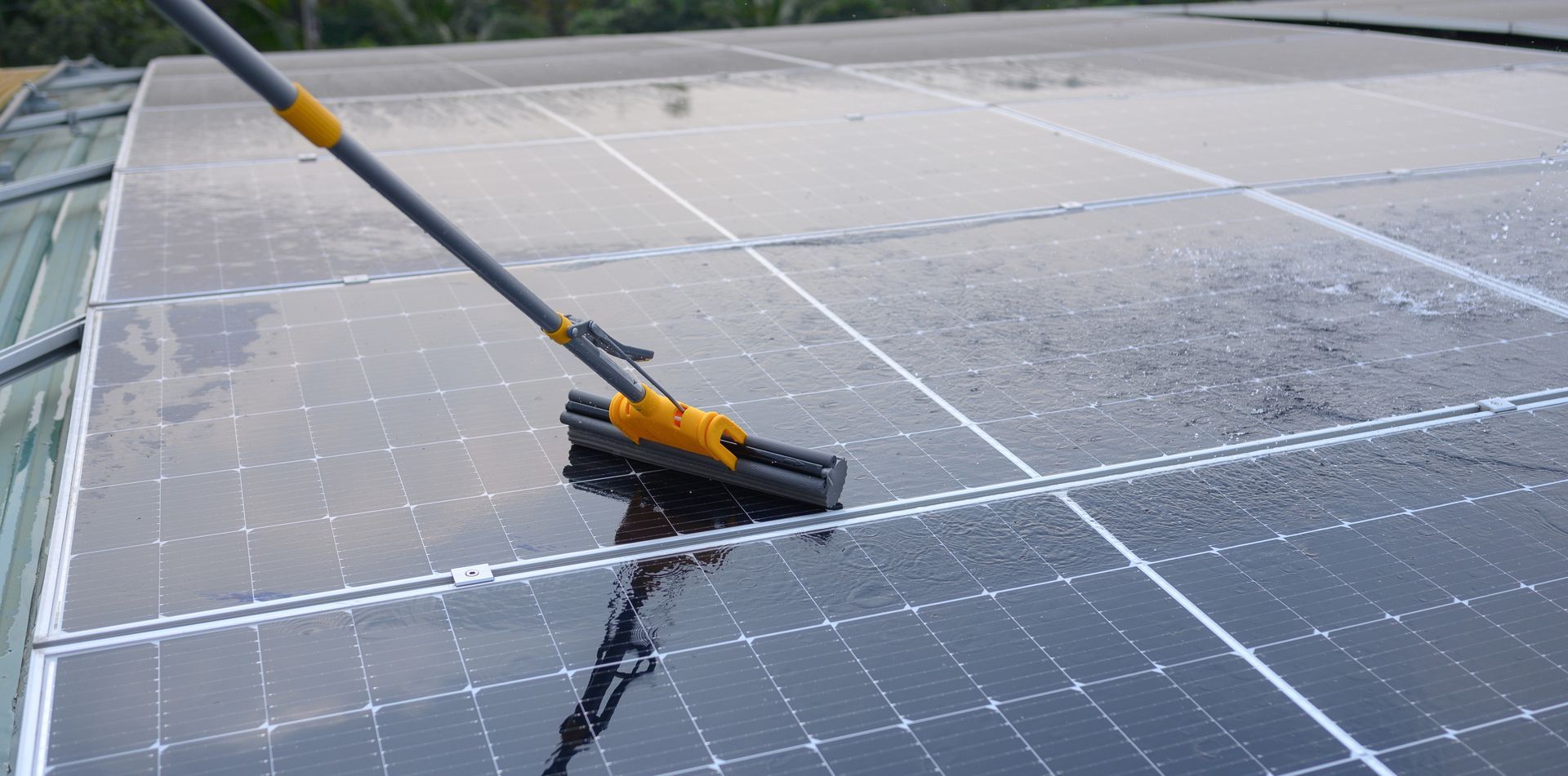 A long-handled squeegee cleans a section of dirty solar panels on a roof, revealing a clean, dark surface.