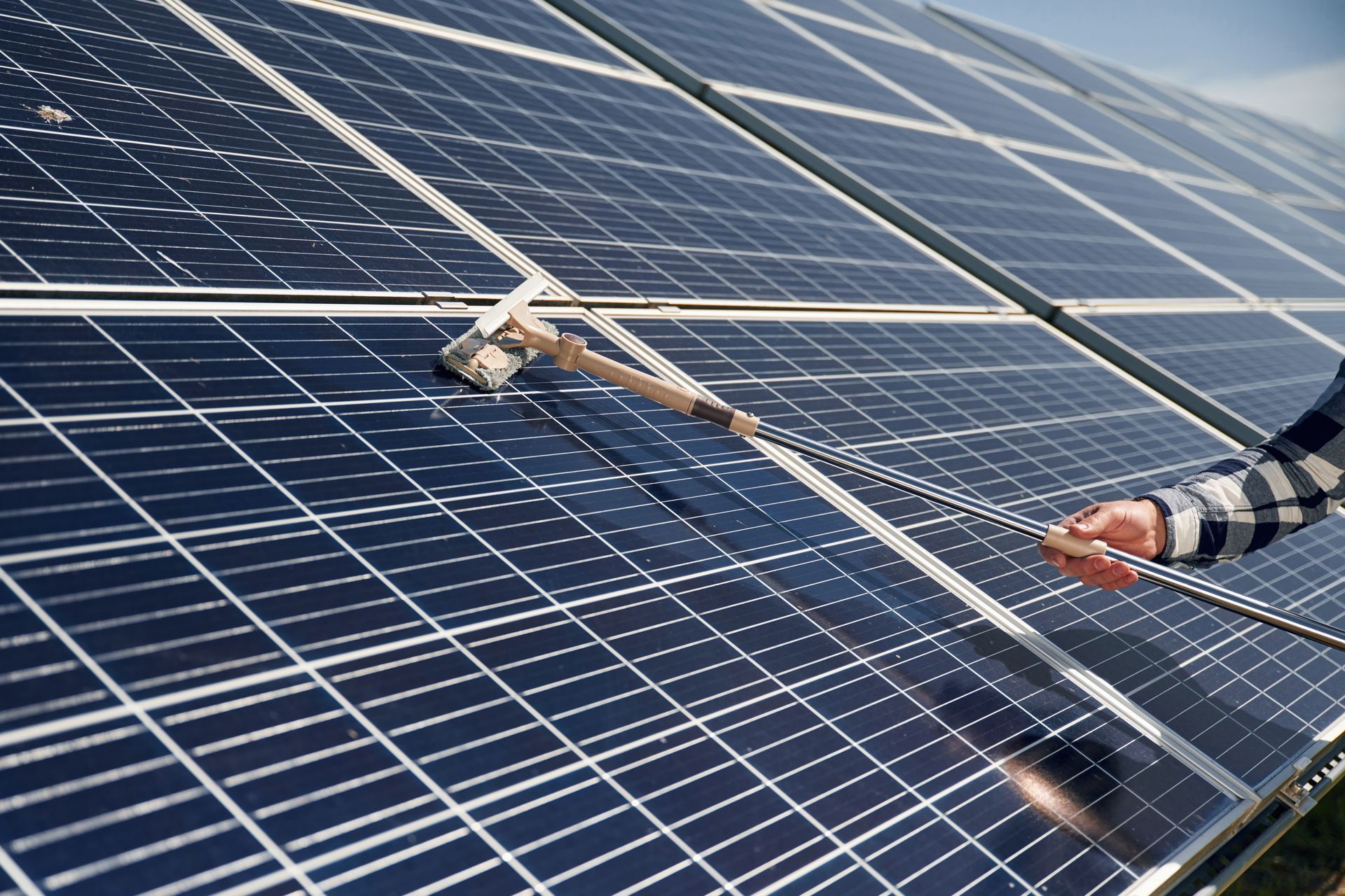A person cleans the surface of a solar panel using a long-handled squeegee.