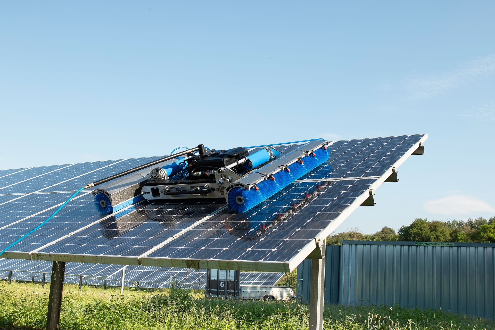 A blue autonomous cleaning robot on tracks moves across a slanted, dark solar panel in a sunny outdoor field.