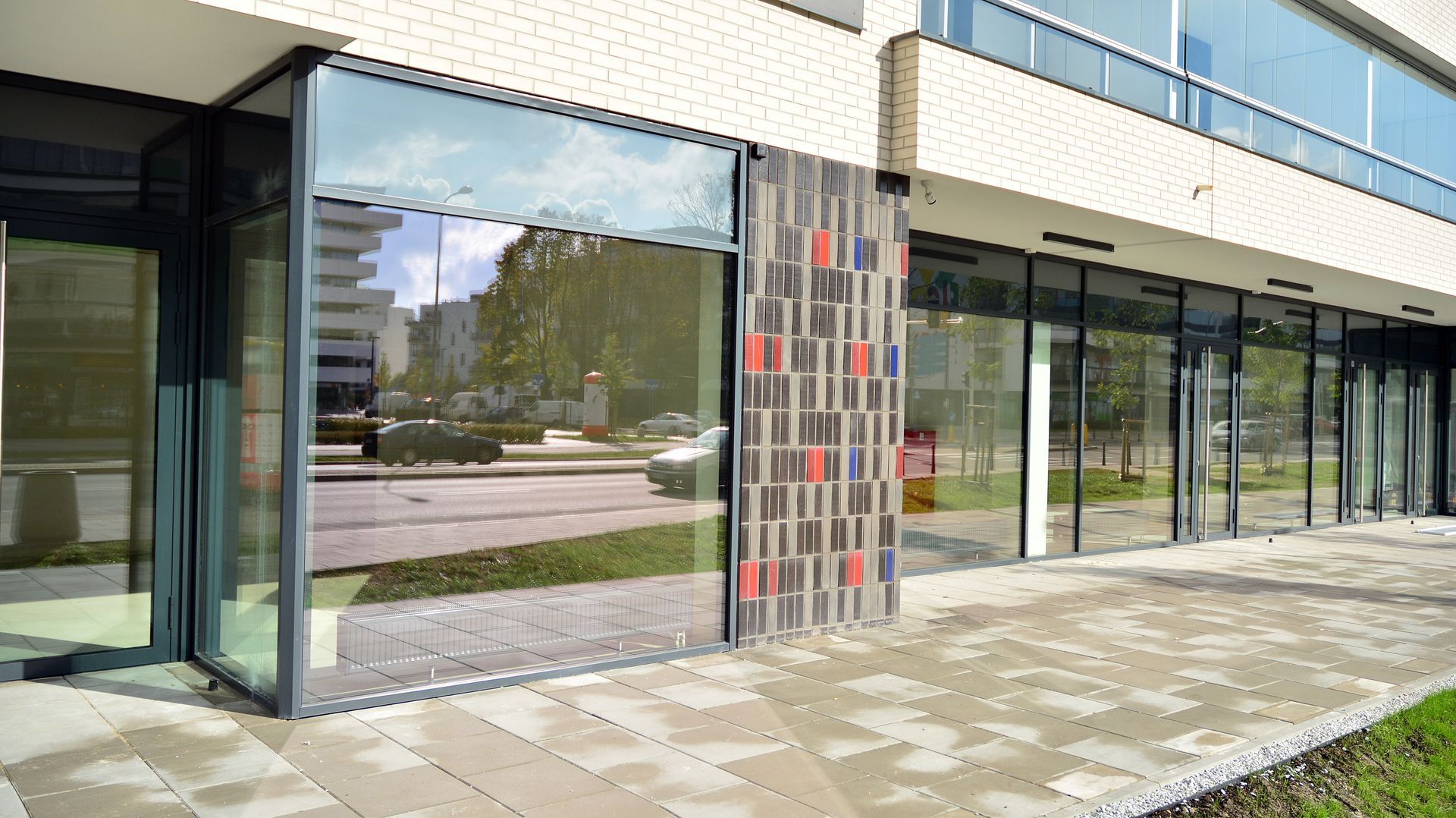 A street-level view of a modern building with large glass windows, a decorative tile panel, and a paved walkway in front.