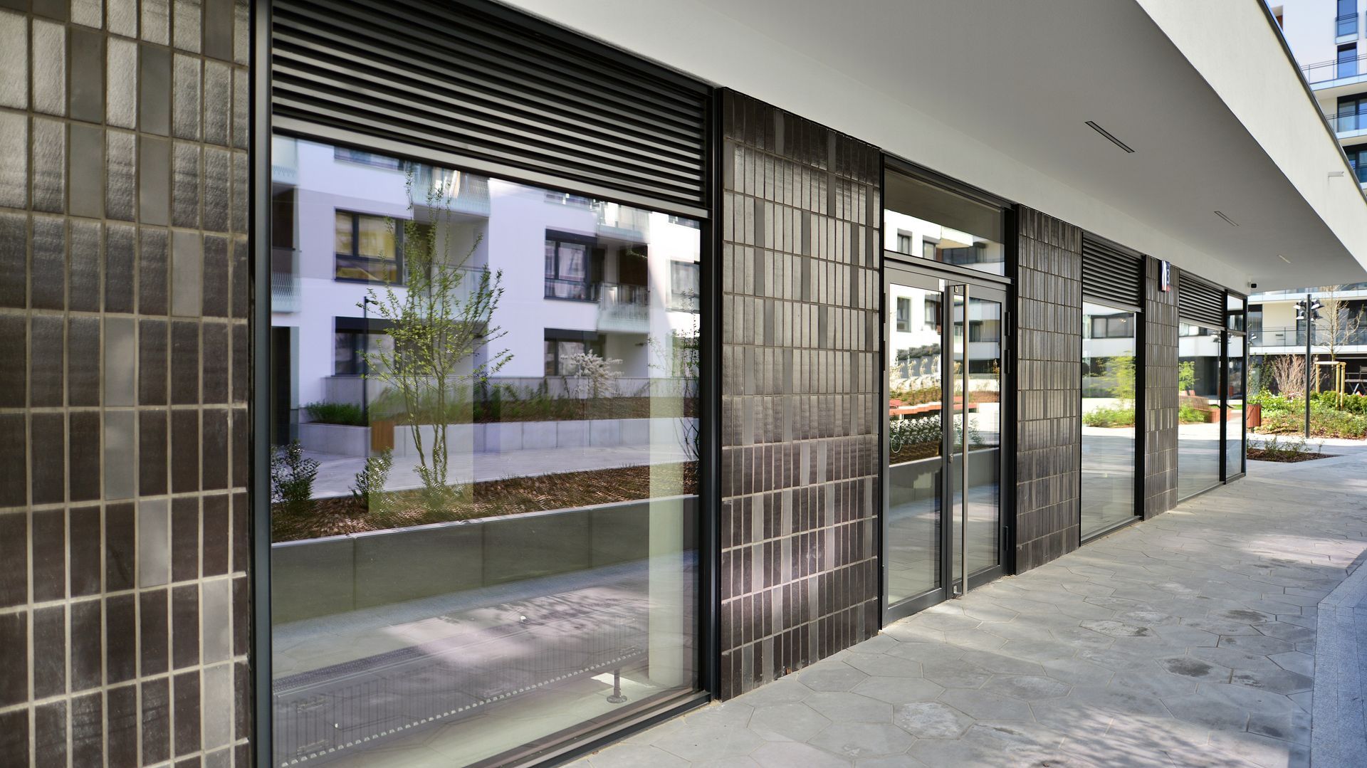 Modern storefront with dark vertical tiles, large glass windows, and metal shutters under a minimalist overhang.