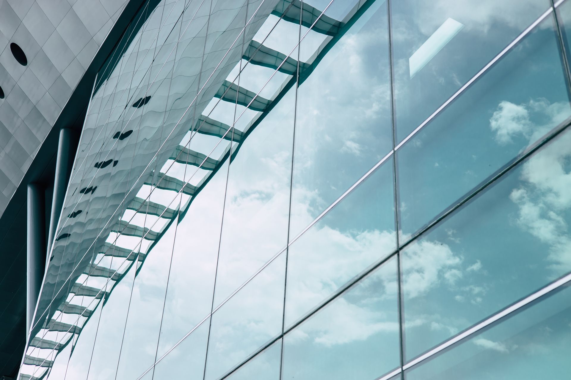 A low-angle view of a modern glass building featuring a prominent metallic, sloped roof reflecting a cloudy blue sky.