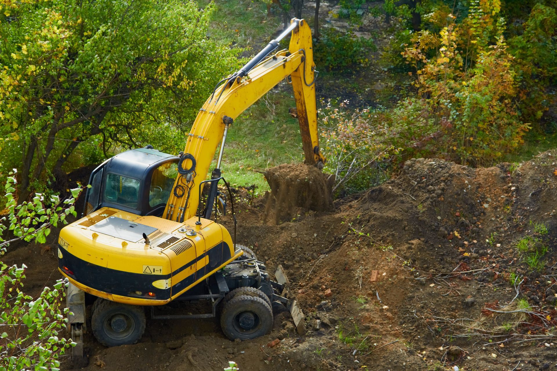 A yellow excavator sits in a dirt-filled, wooded area, digging into the earth with its extended mechanical arm.