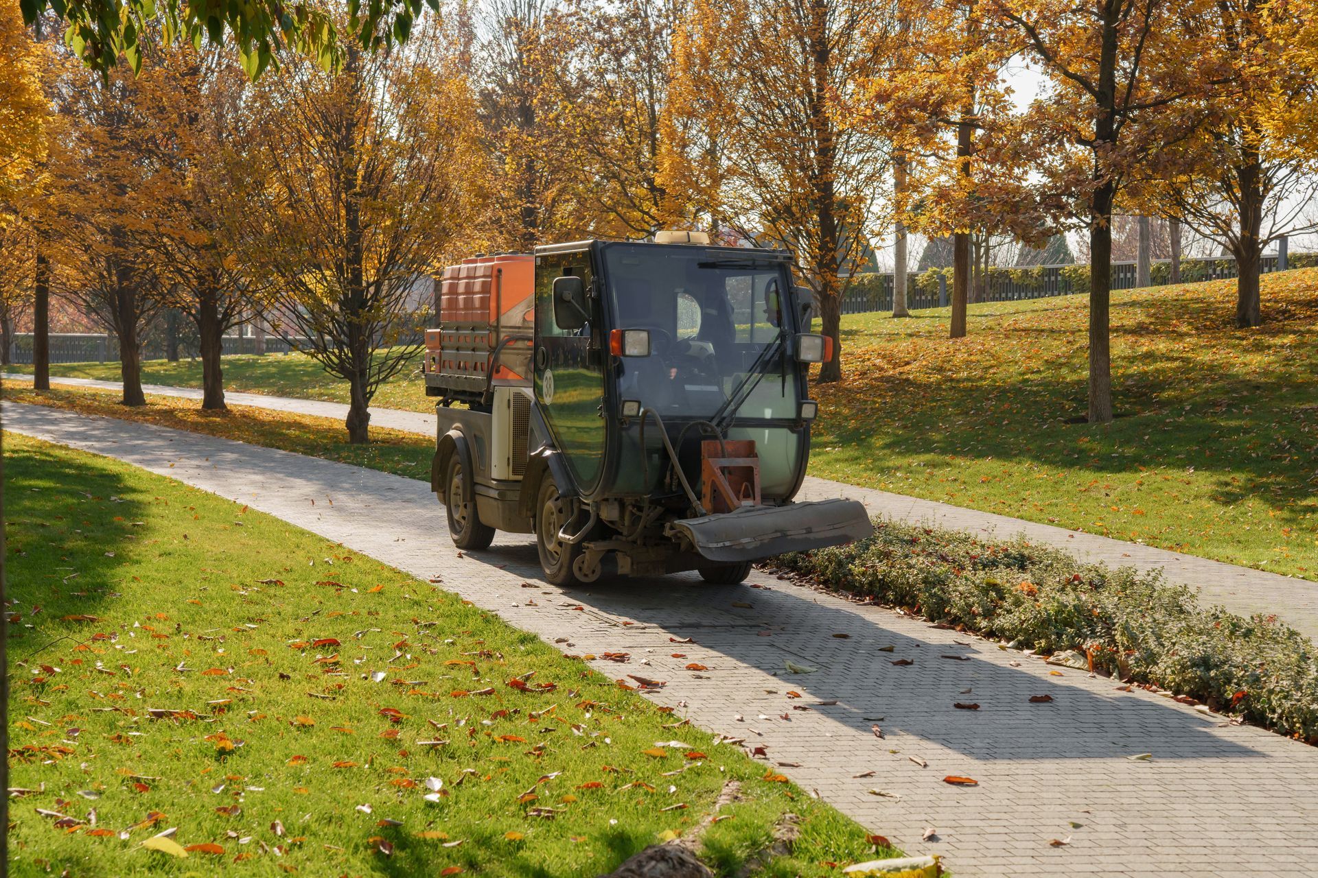 A small, orange and black street sweeper clearing leaves from a paved path in a park during autumn.