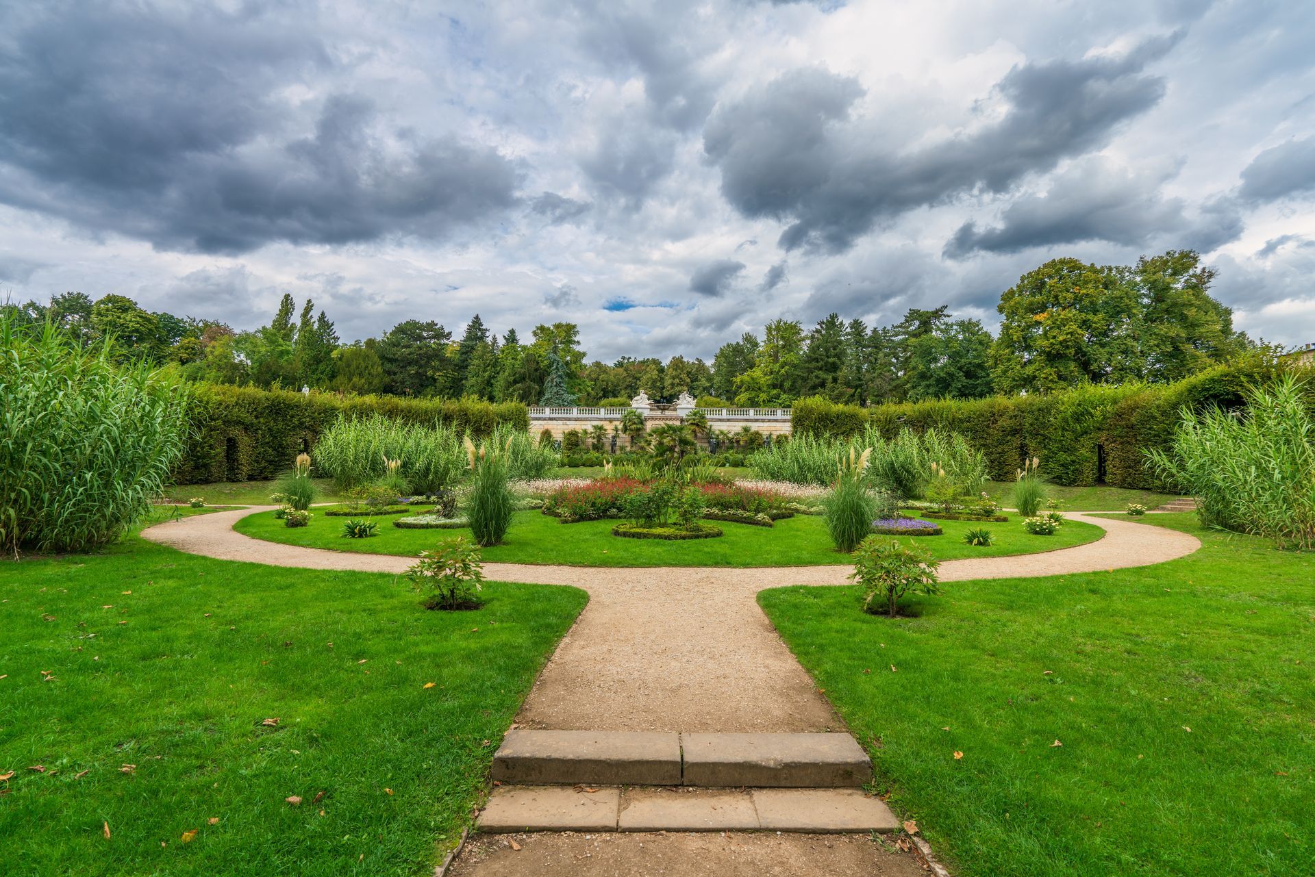A gravel path leads to a circular flower garden in a lush park under a dramatic, cloudy sky.