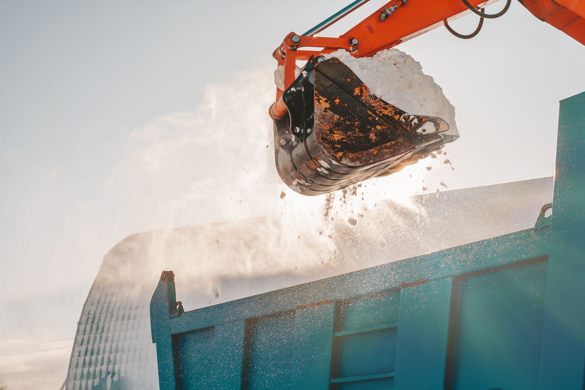 An orange excavator bucket dumps a load of snow into a large blue truck container against a bright, hazy sky.