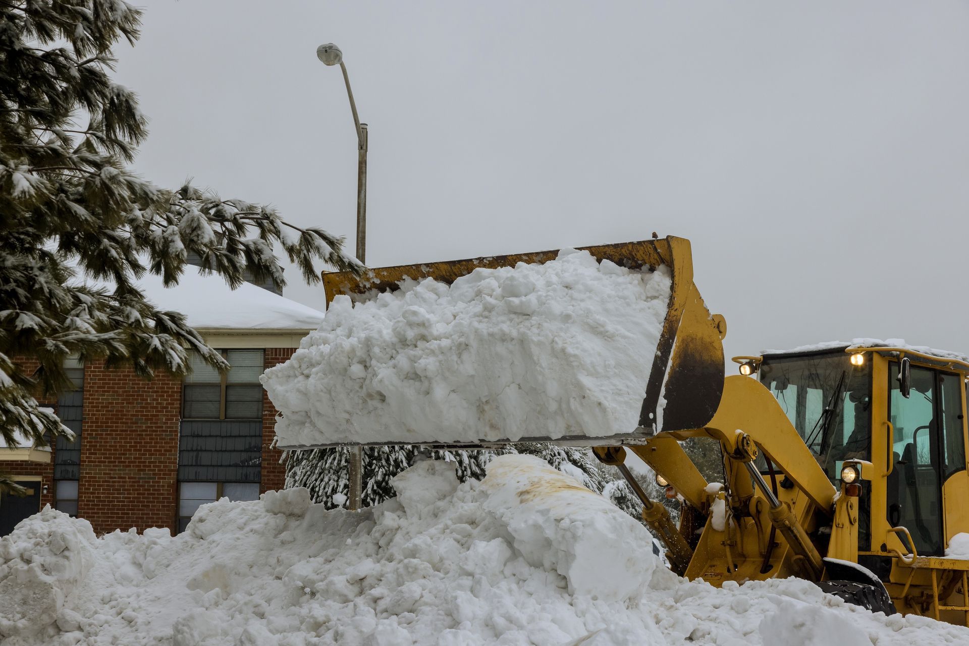A yellow front-end loader clears a large pile of snow from a parking area near a brick building.