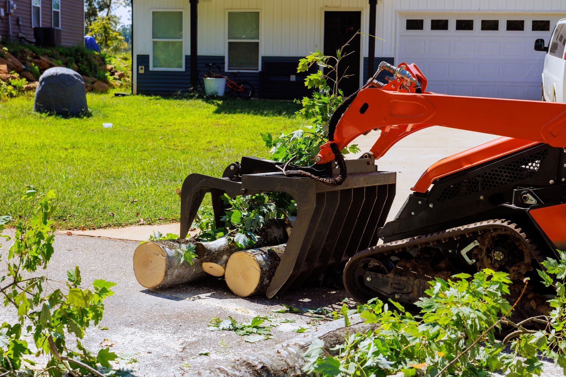 An orange skid steer loader with a grapple attachment moves cut logs and tree branches in a residential driveway.