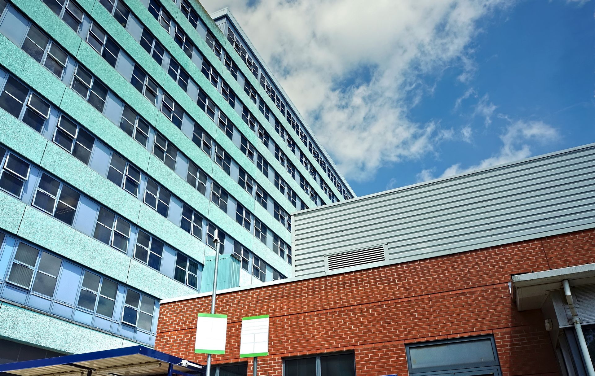 A tall, light-blue building towers over a shorter, red-brick structure against a bright, cloudy blue sky.